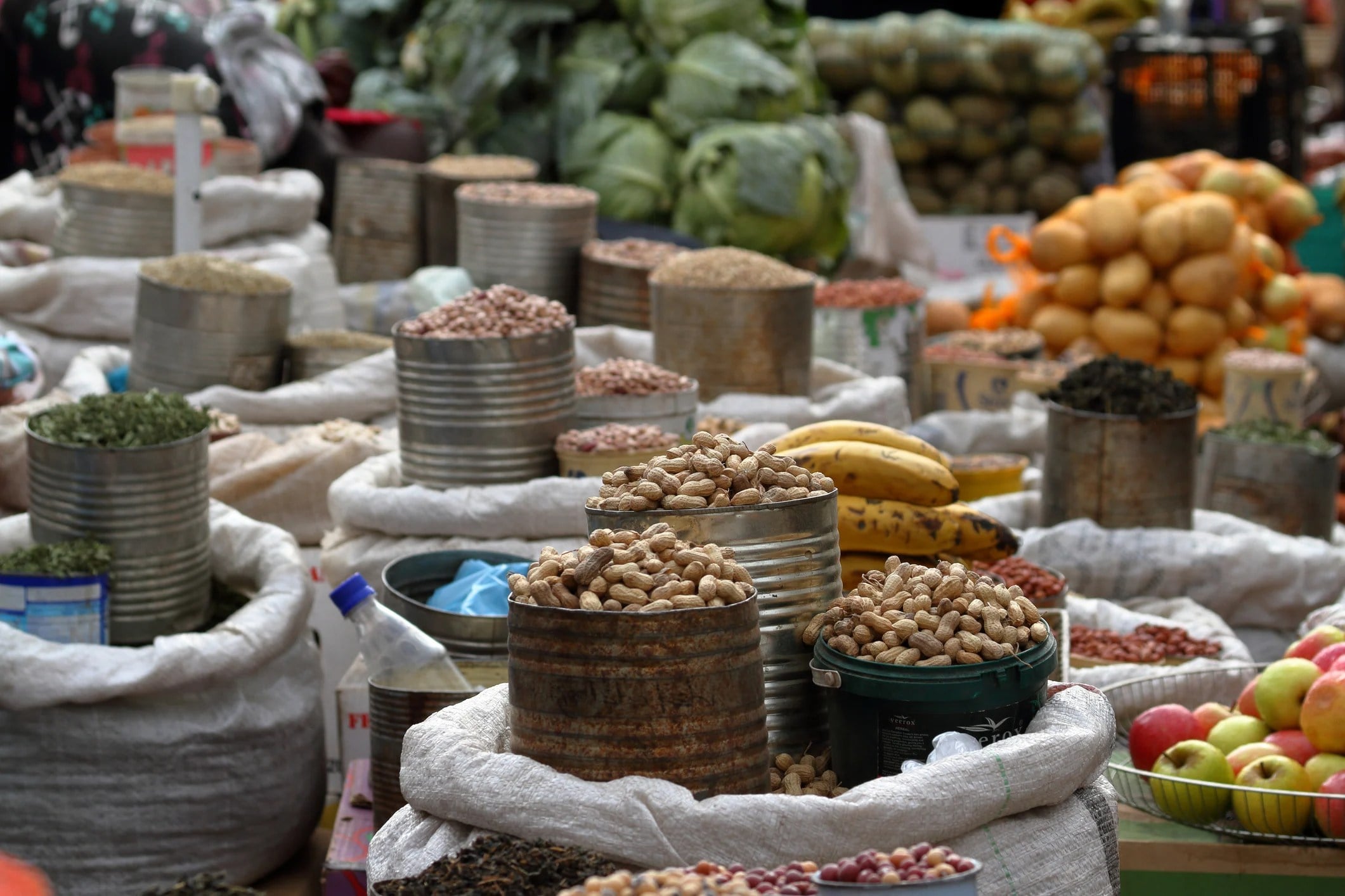Various sacks of nuts and legumes are arranged in a bustling market, with fruits and vegetables in the background.