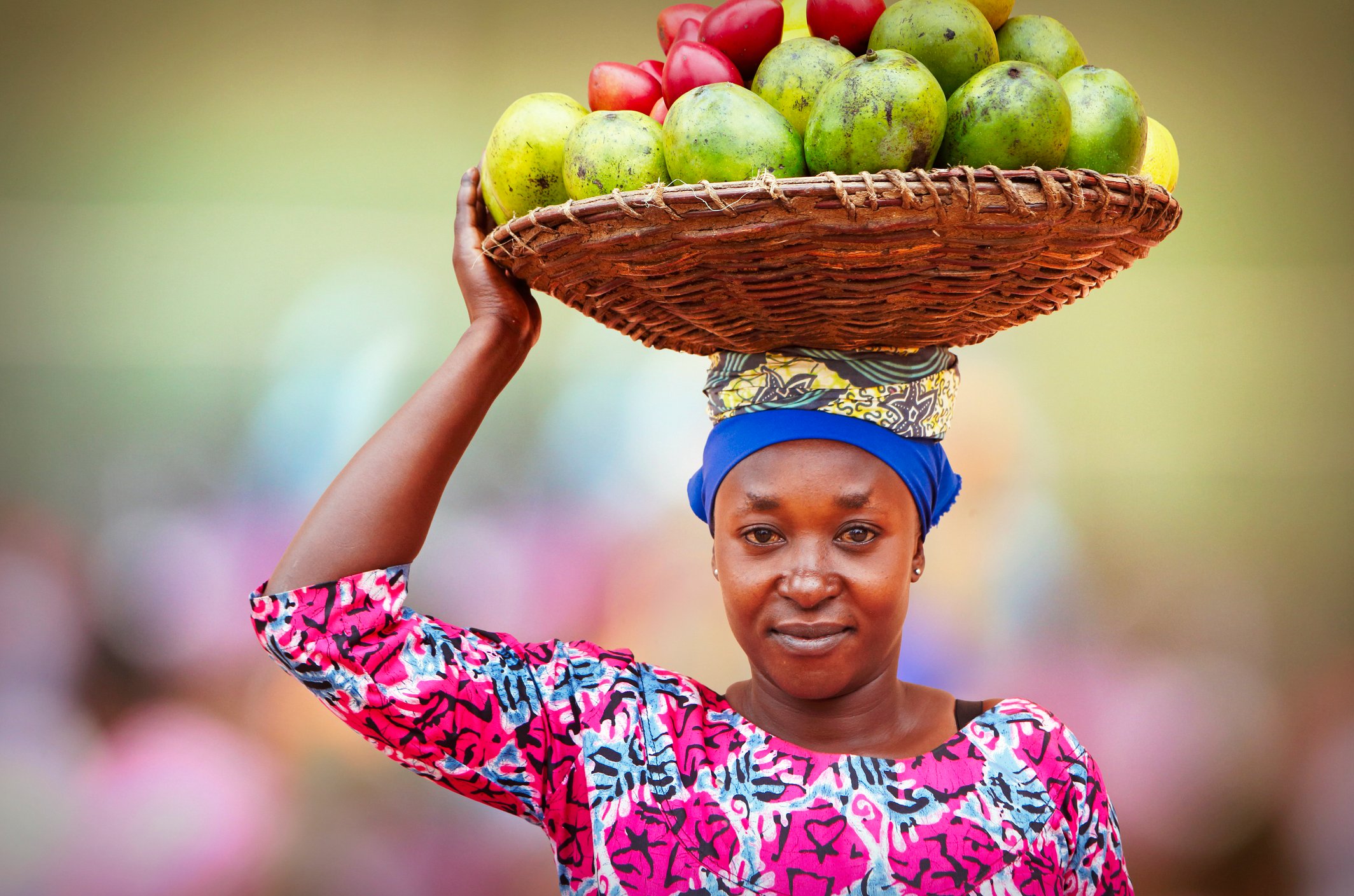 A woman in a colorful outfit balances a basket of fruits on her head while smiling at the camera.