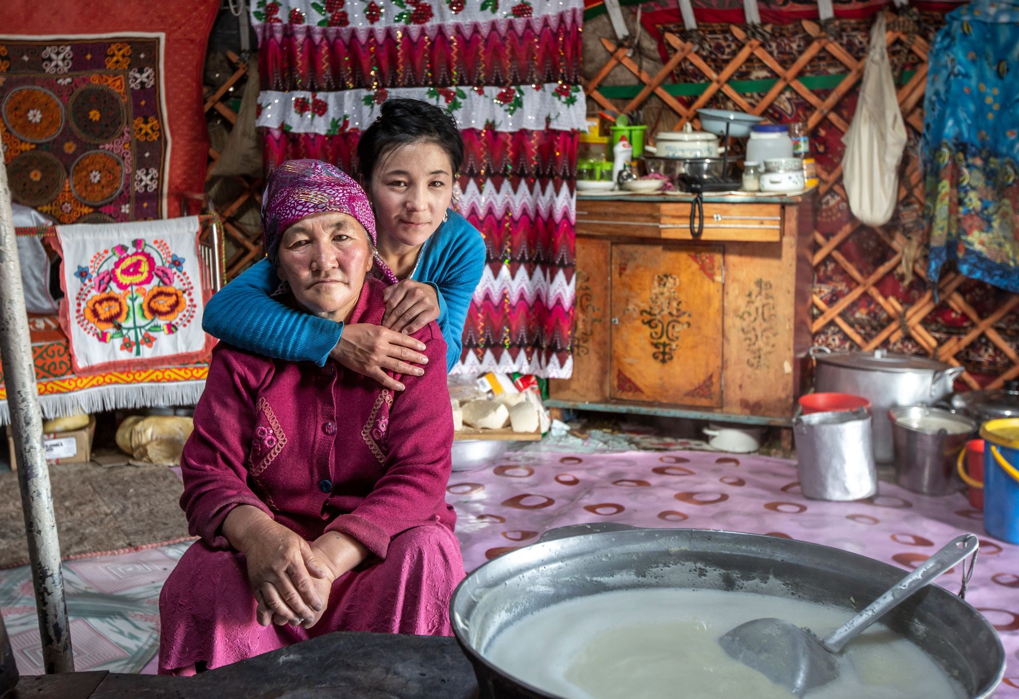 A young girl embraces an elderly woman in a cozy, traditional yurt adorned with colorful textiles and kitchen items.