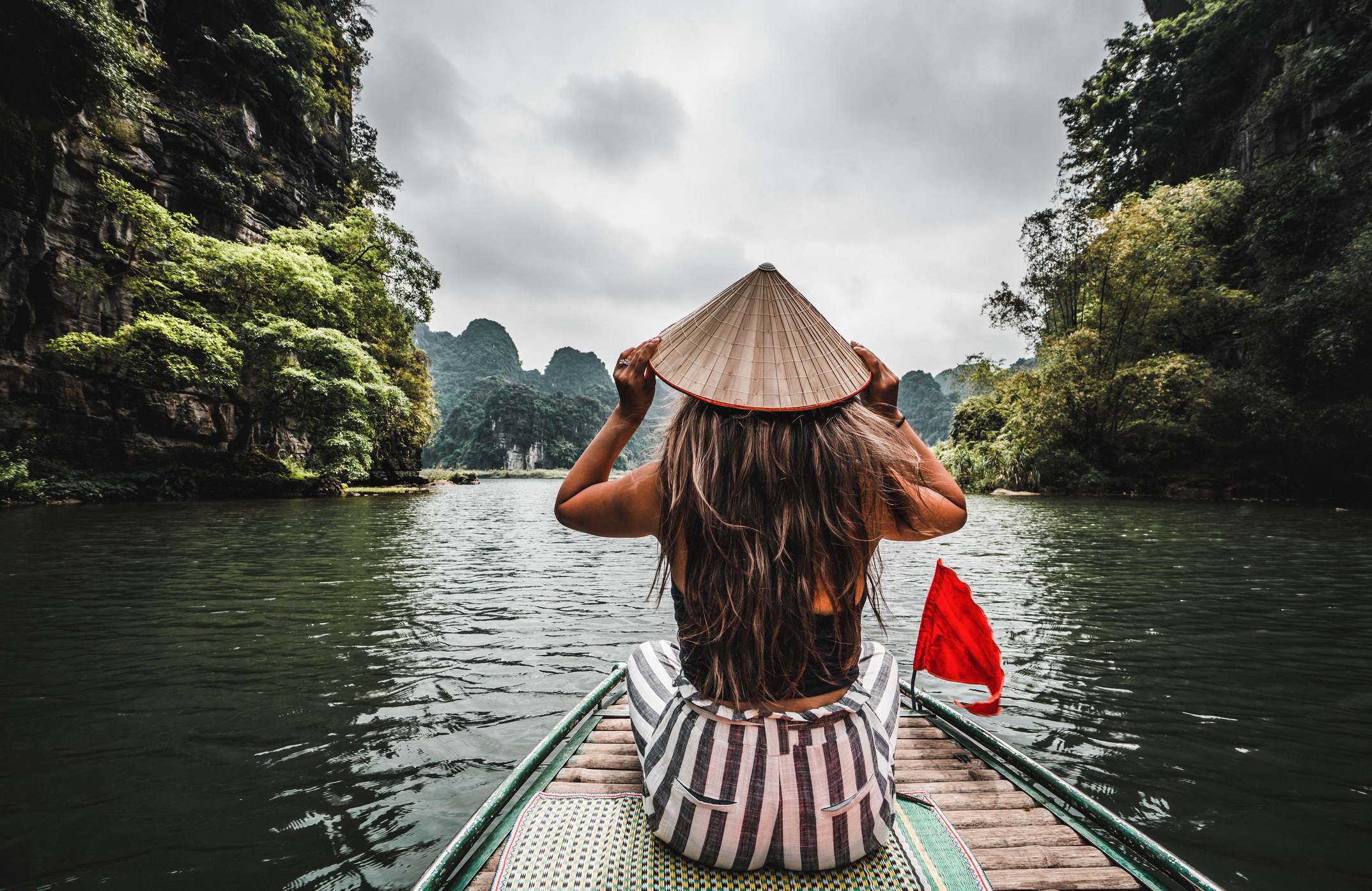 A person in a conical hat sits on a boat, gazing at scenic mountains and lush greenery on a calm lake.