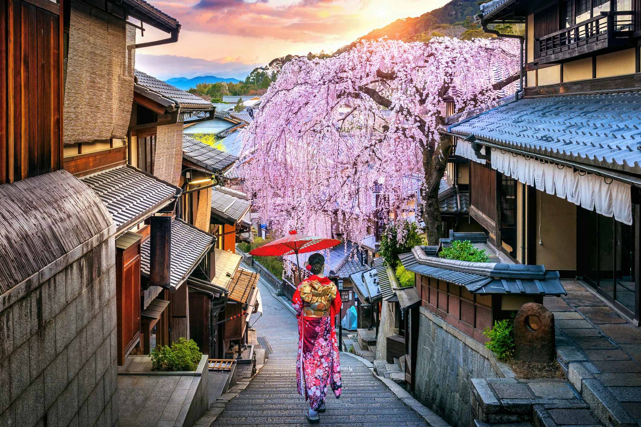 A person in a colorful kimono walks down a historic street with cherry blossoms and a sunset in the background.