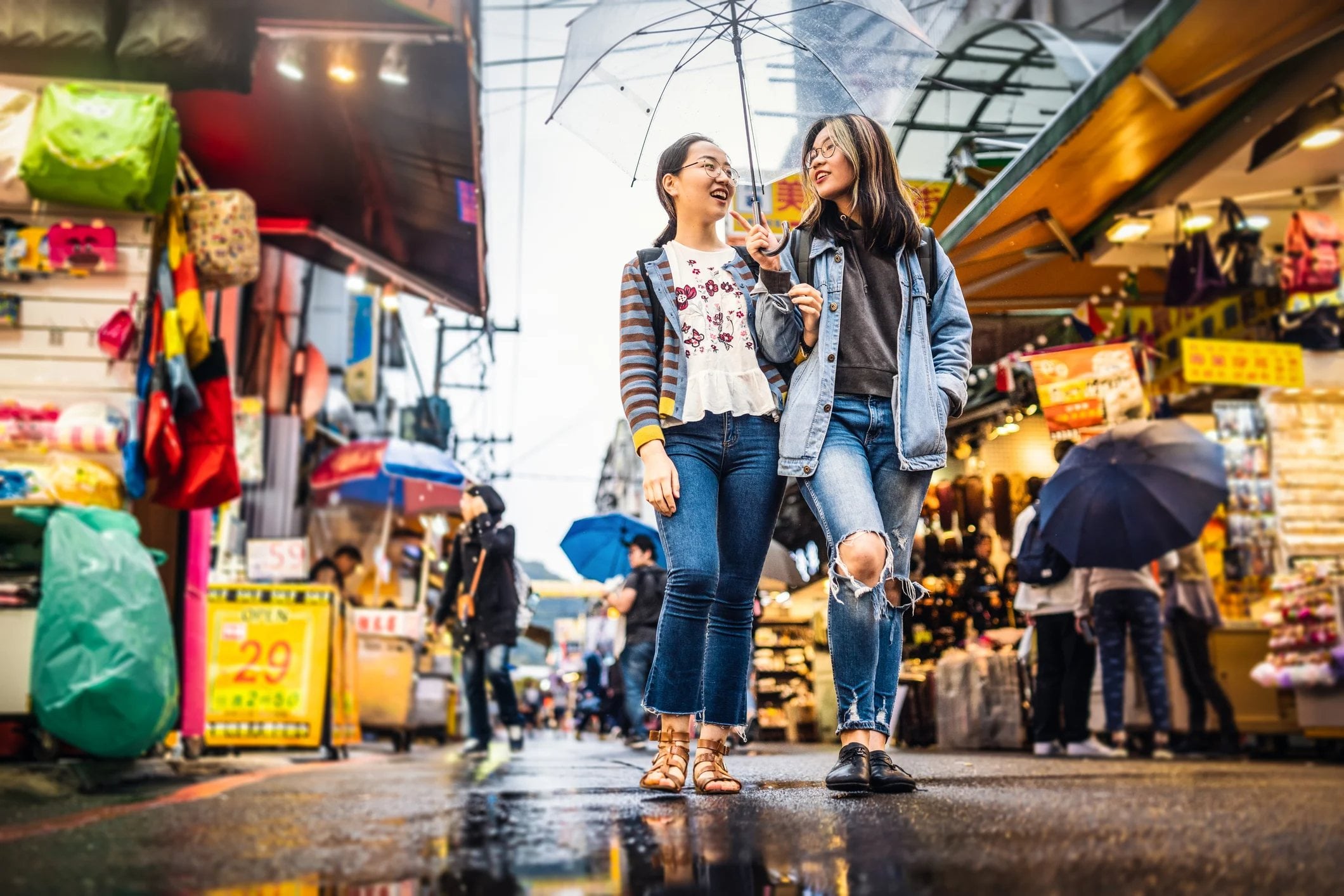 Two friends walk together under an umbrella in a vibrant market, surrounded by colorful stalls and wet pavement.