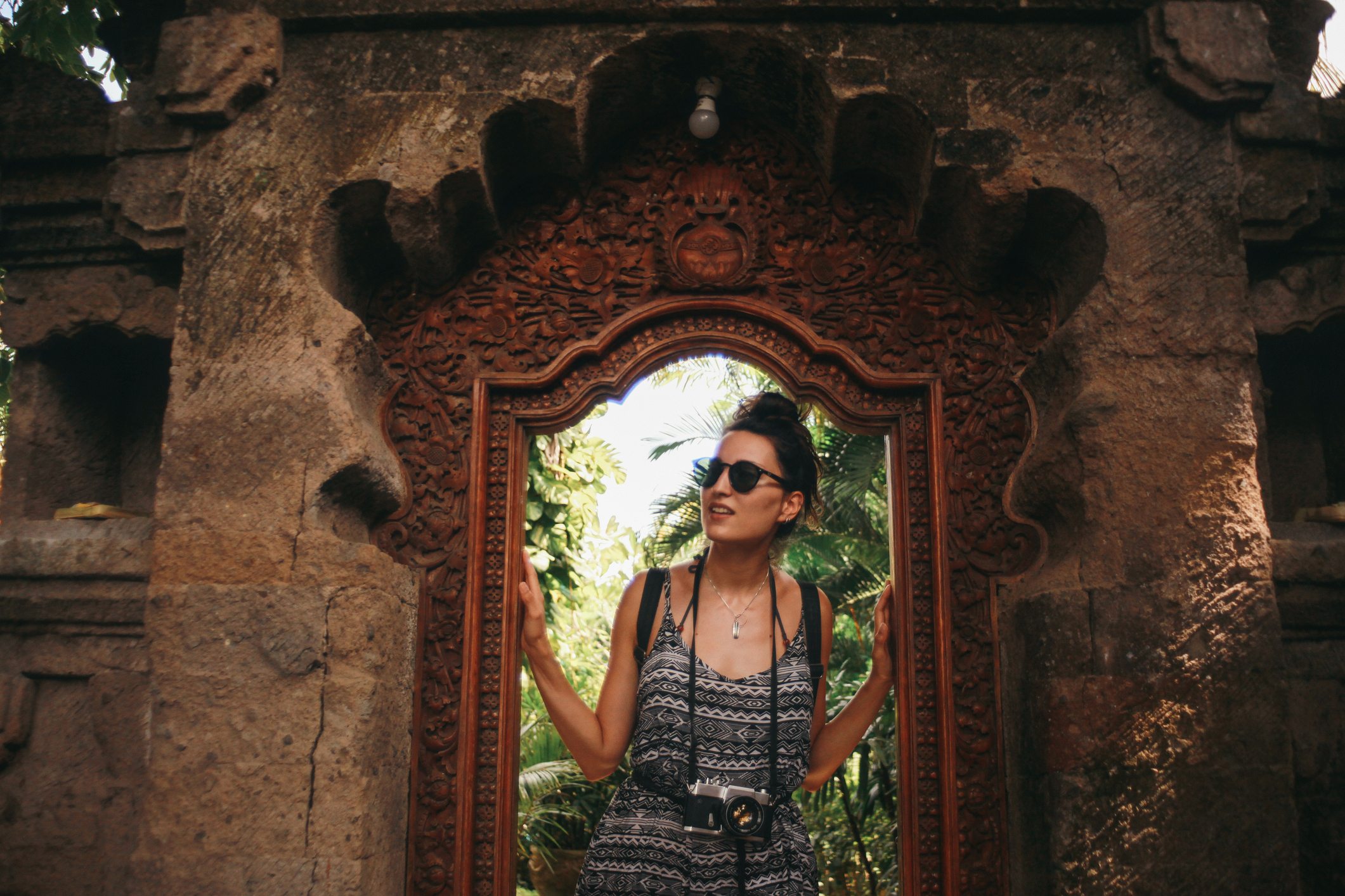 A person in sunglasses stands in a carved stone doorway, surrounded by greenery. A camera hangs from their neck.