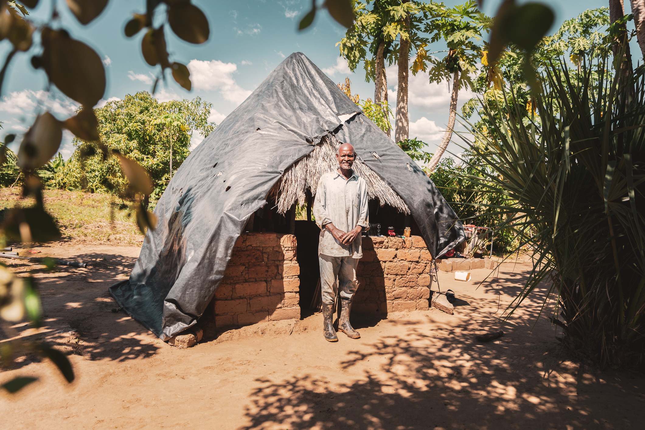Man standing in front of a small mud brick house with a thatched roof in a natural outdoor setting.
