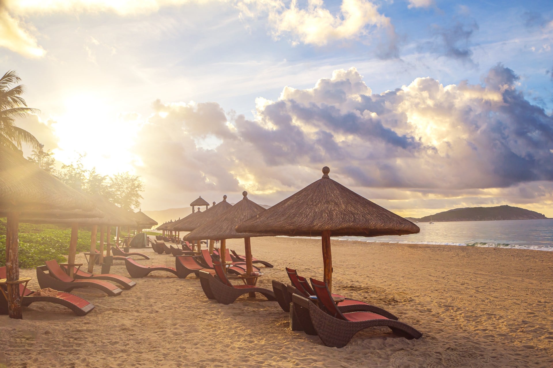 Beach scene with lounge chairs under straw umbrellas, illuminated by a warm sunset and scattered clouds.
