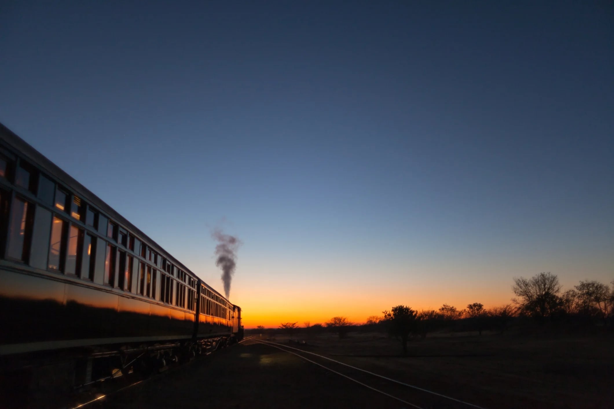 A steam train travels along tracks at sunset, with vibrant colors in the sky and a silhouette of trees in the distance.