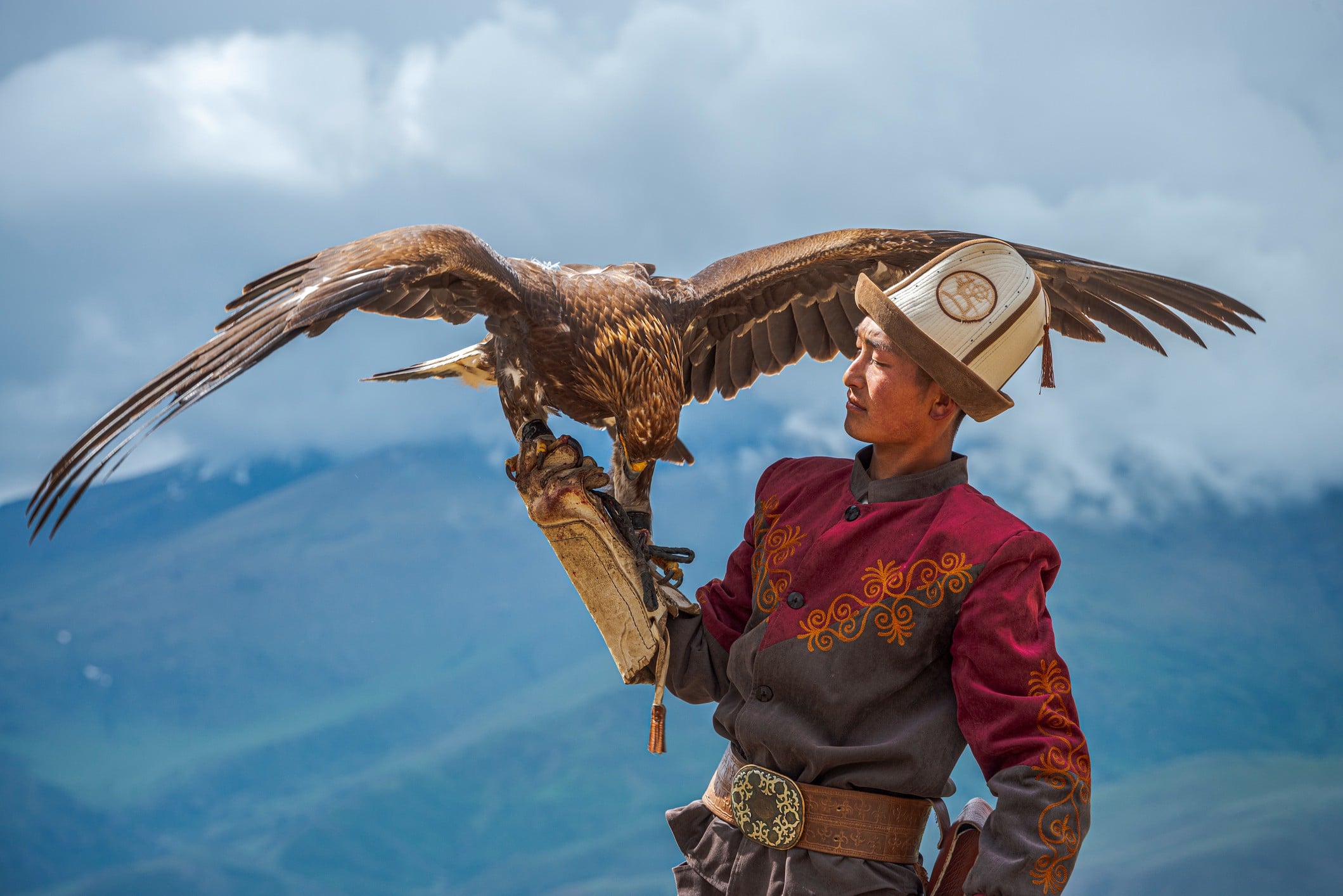 A man in traditional attire holds a large eagle on his arm against a mountainous backdrop.