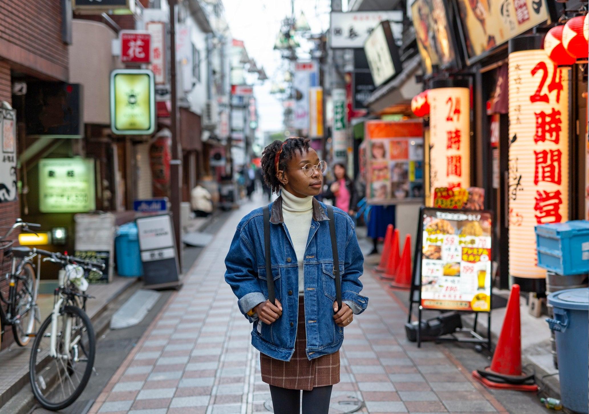 A person in a denim jacket stands in a vibrant urban alley filled with shops and neon signs.