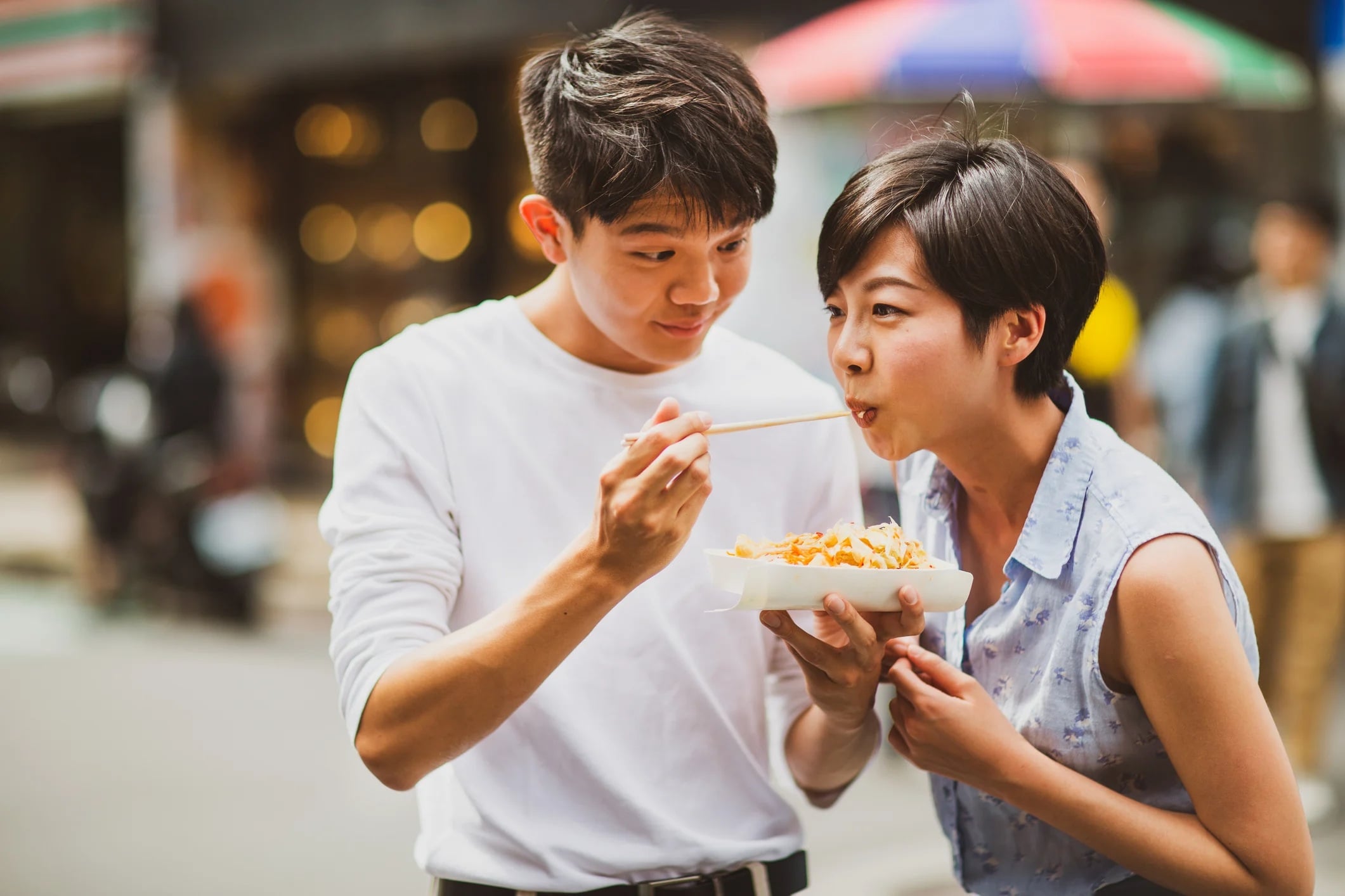 A young man feeding a young woman noodles on a bustling street, both smiling and enjoying the moment.