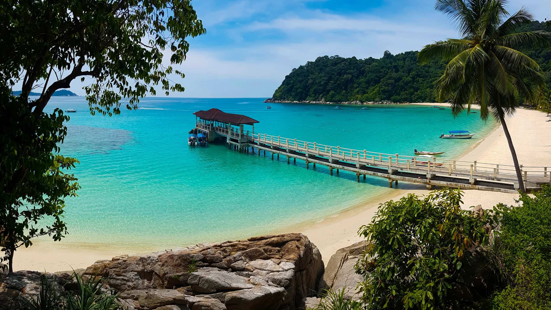 A scenic beach with a wooden pier extending into turquoise waters, surrounded by lush greenery and mountains in the background.