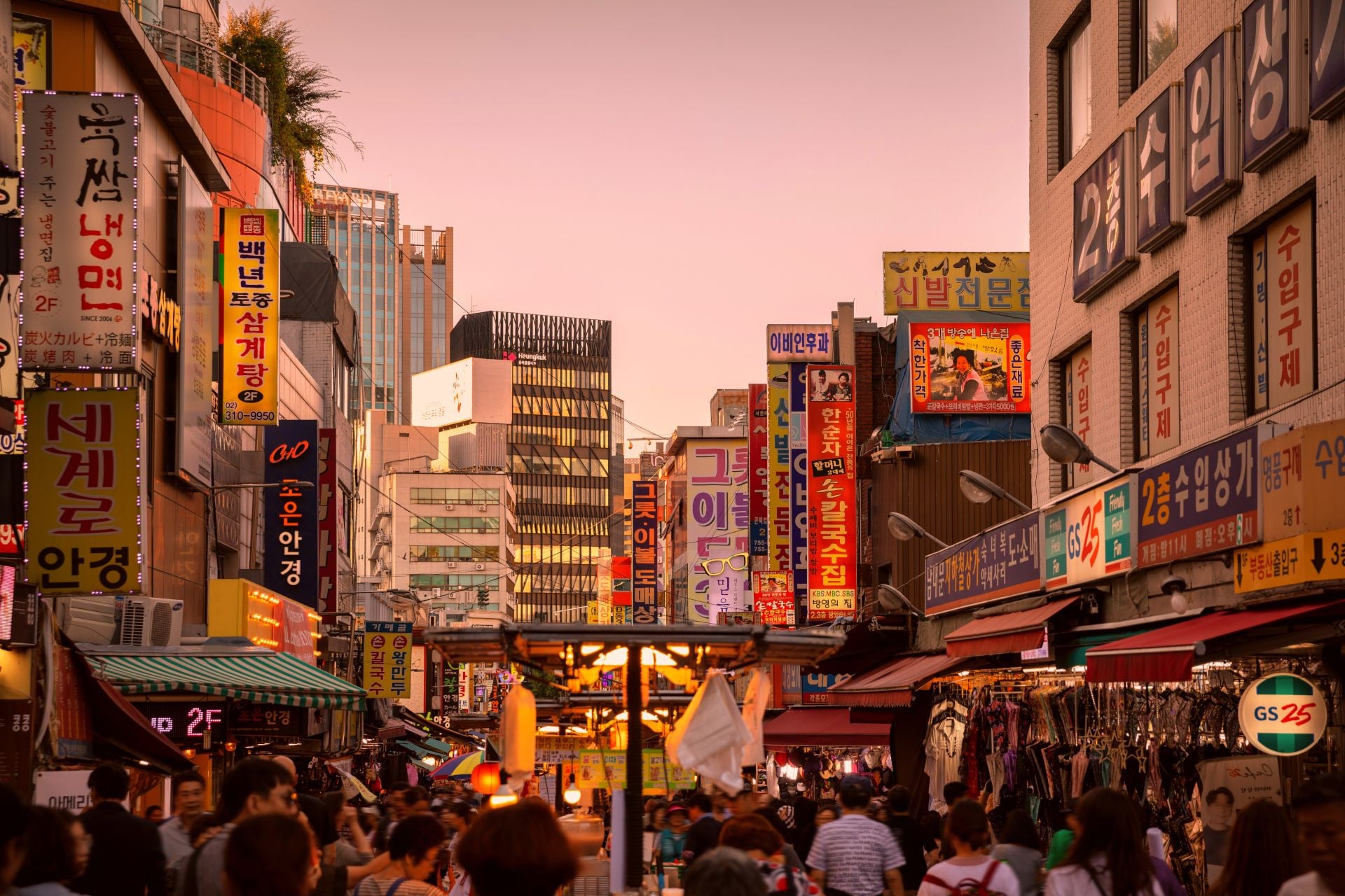 A bustling street market at dusk, filled with colorful signs and people shopping among stalls.
