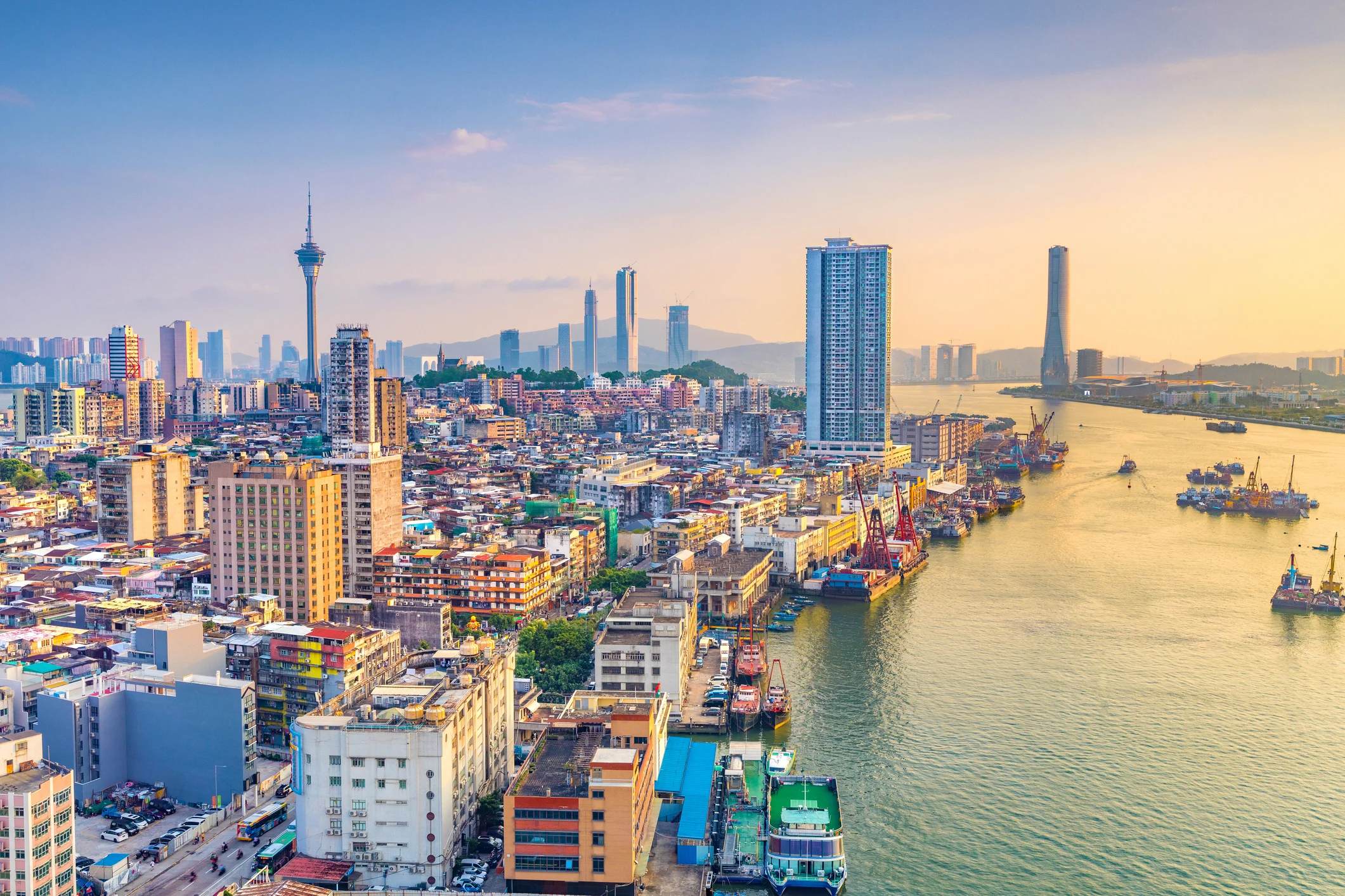 A scenic cityscape at sunset, featuring a river, skyscrapers, and boats along the waterfront.