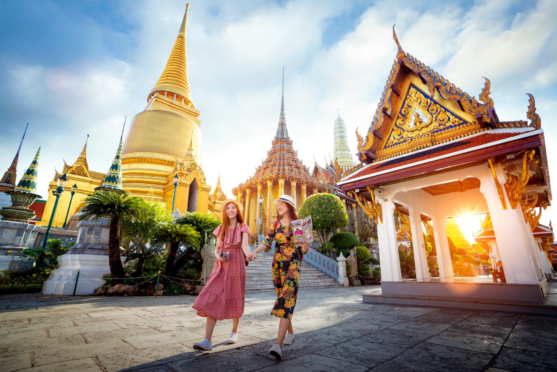 Two smiling women walk hand in hand, surrounded by ornate temples and golden spires, under a bright sky at sunset.