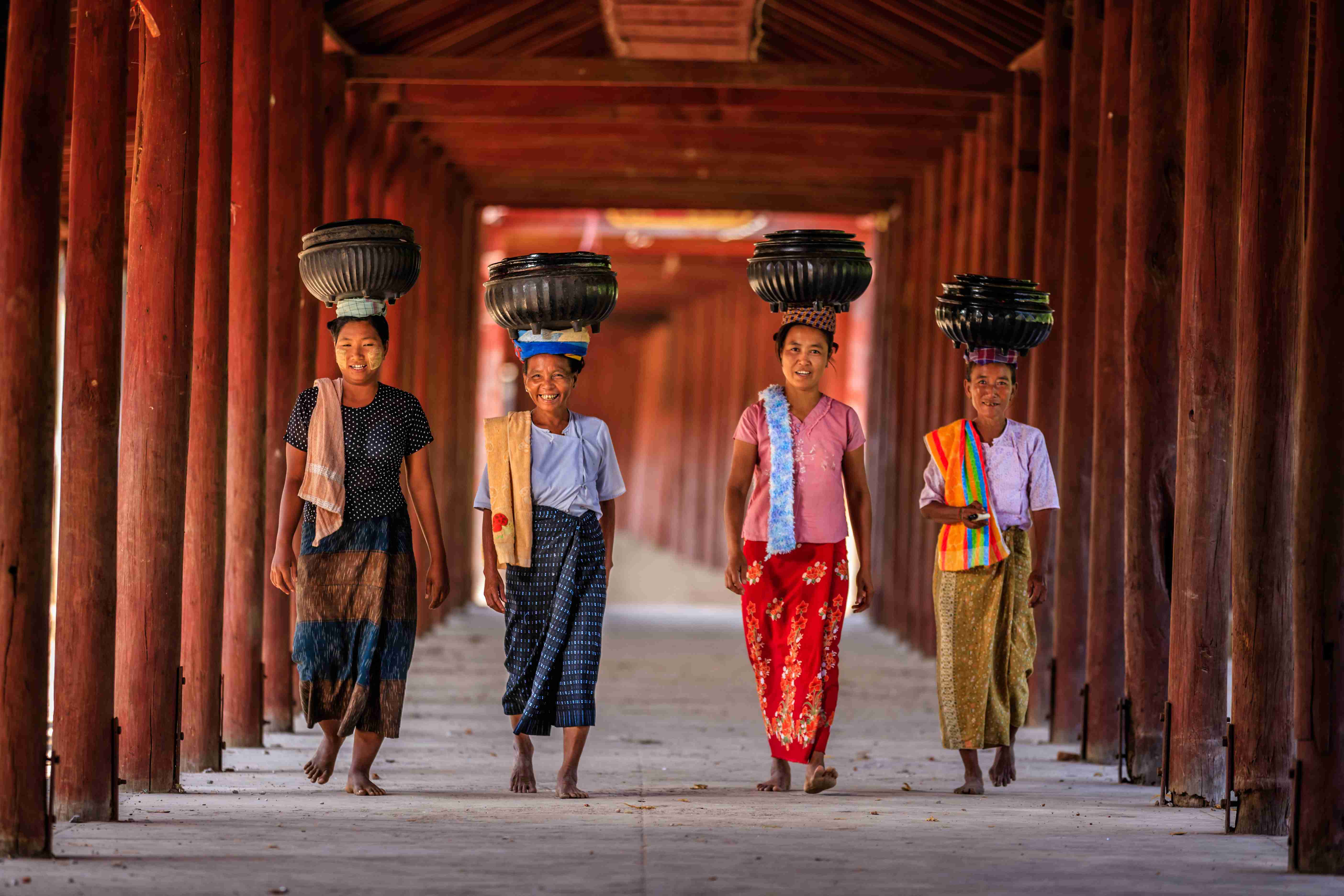 Four girls walk confidently under a wooden archway, each balancing a container on their heads.