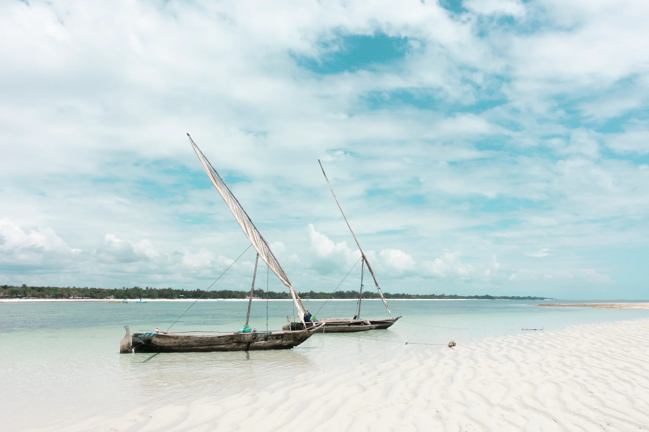 Two traditional sailboats on a calm beach, with soft sand and a cloudy sky in the background.