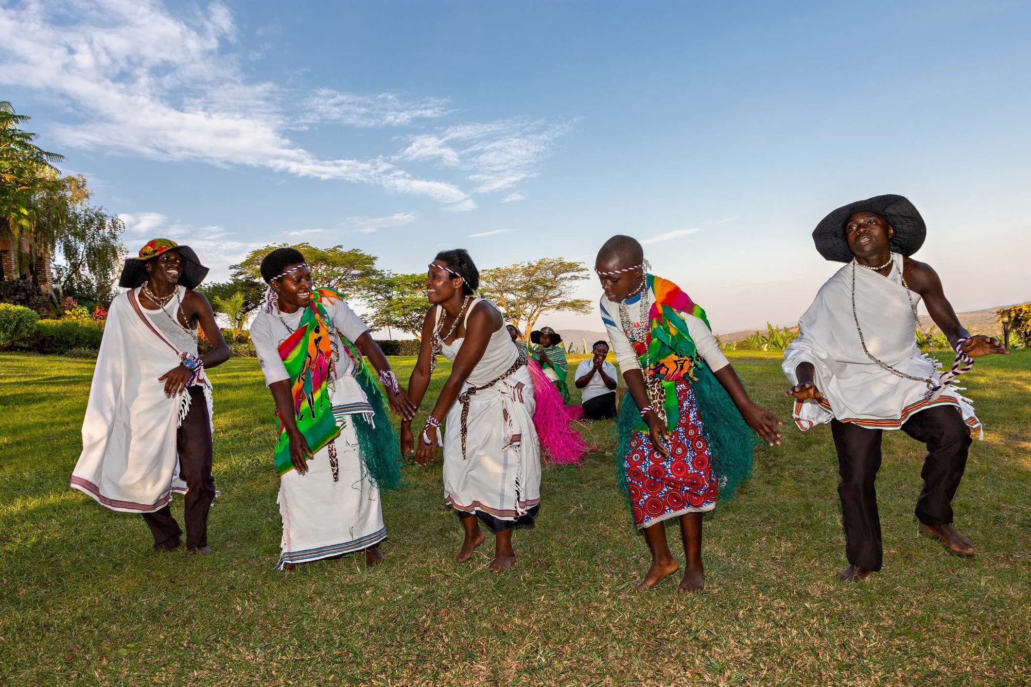 Group of dancers in colorful traditional attire performing outdoors, with a scenic landscape in the background.