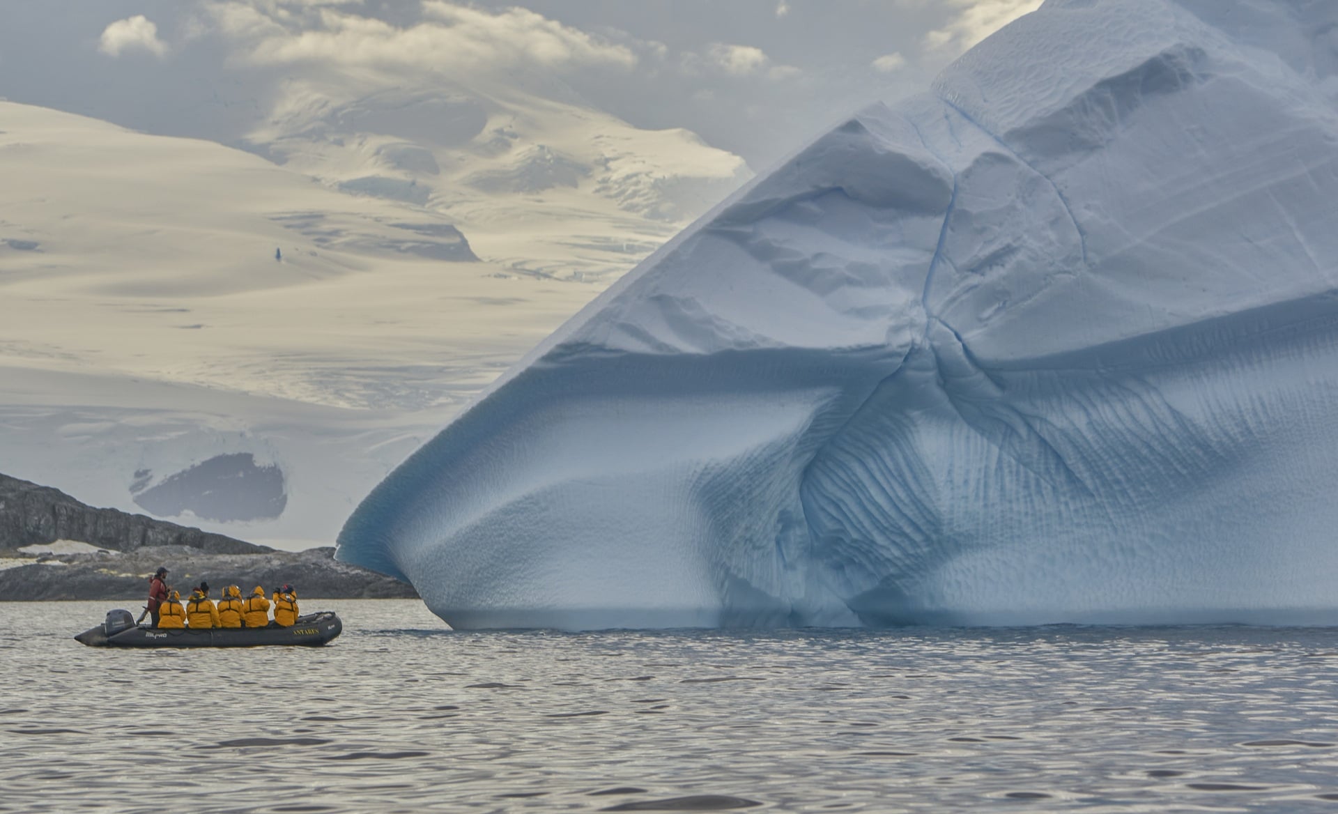 A small boat with passengers approaches a large blue iceberg in a calm sea, surrounded by icy landscapes and clouds.