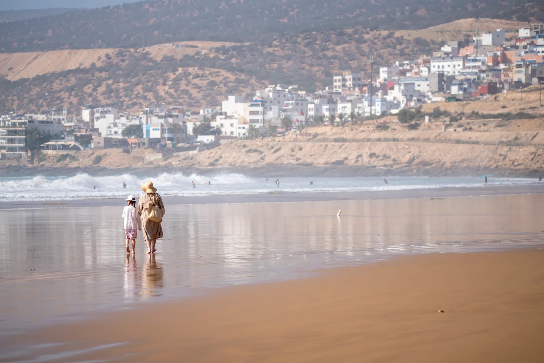 Two people walk along a sandy beach, with waves and a distant coastal town in the background.