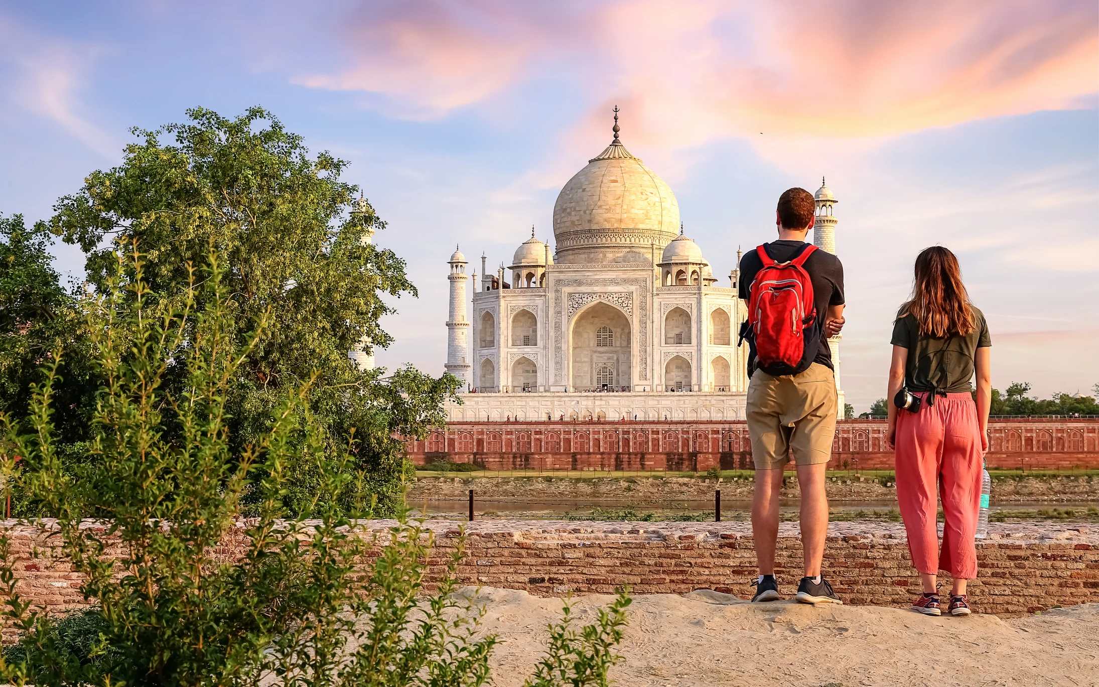 A couple stands on a path, admiring the Taj Mahal against a sunset sky, surrounded by greenery.
