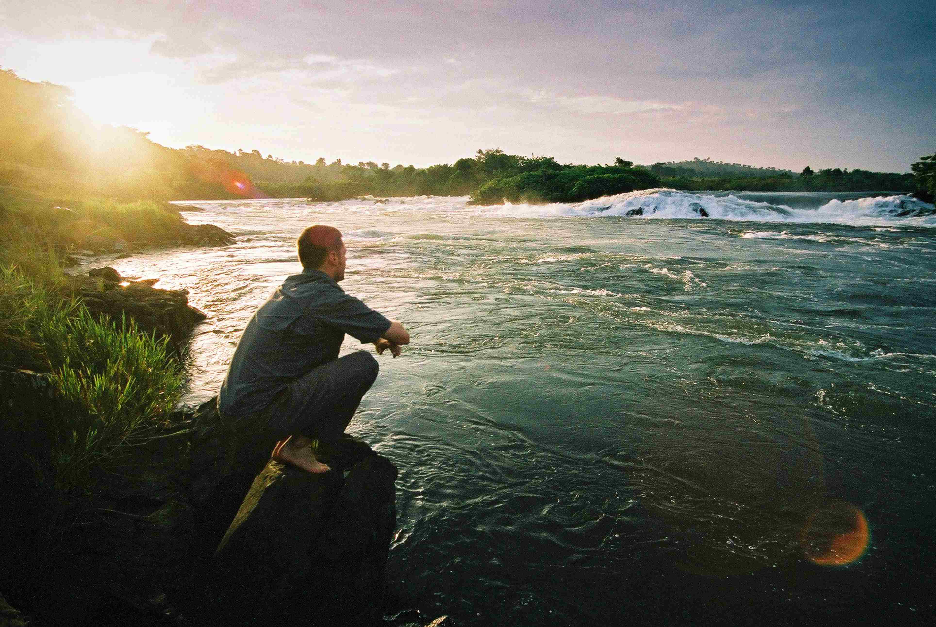 A person sits on a rock by a river, gazing at the water and sunset in a peaceful, natural setting.