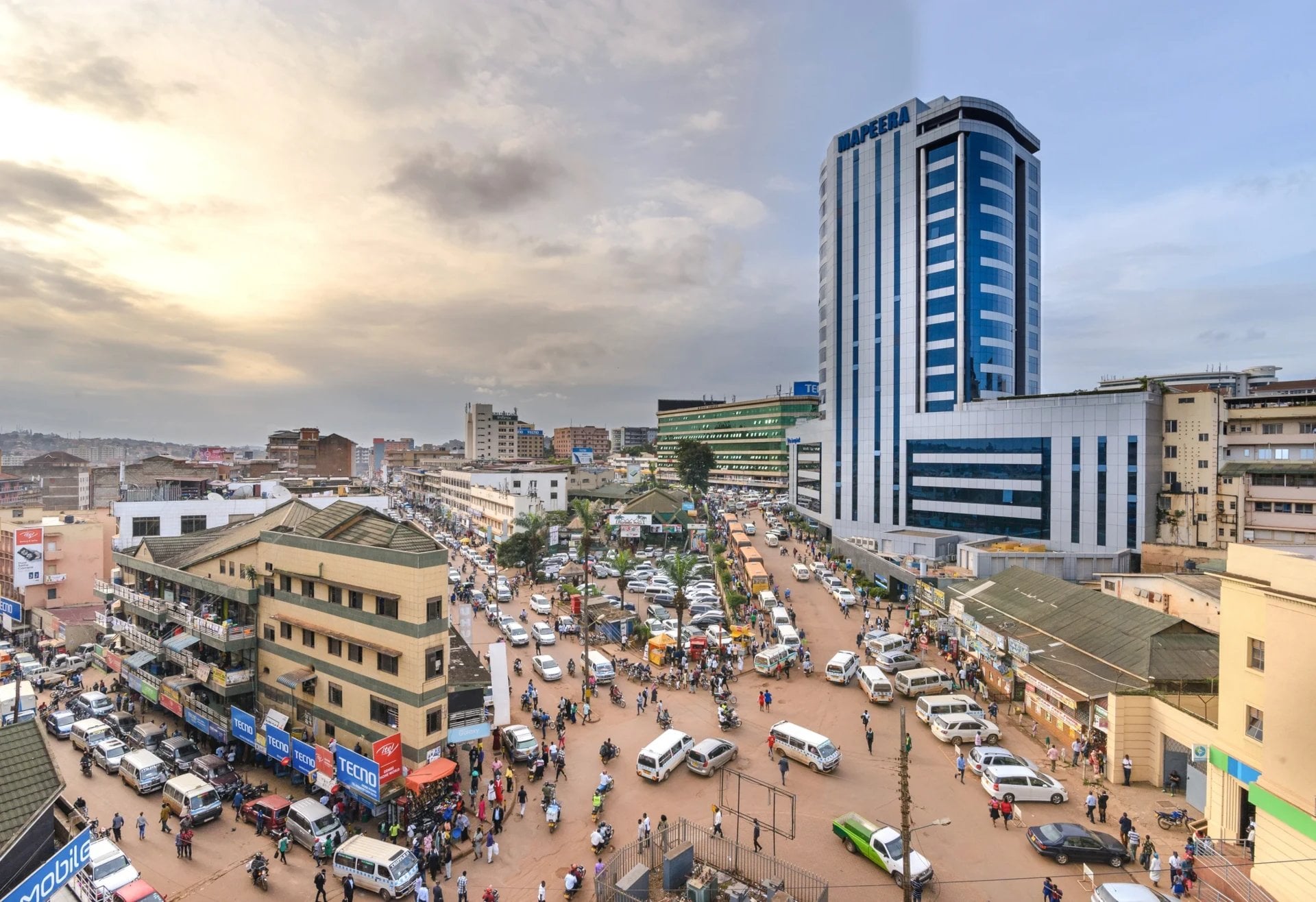 A bustling urban market scene with cars, people, and tall buildings under a cloudy sky during sunset.