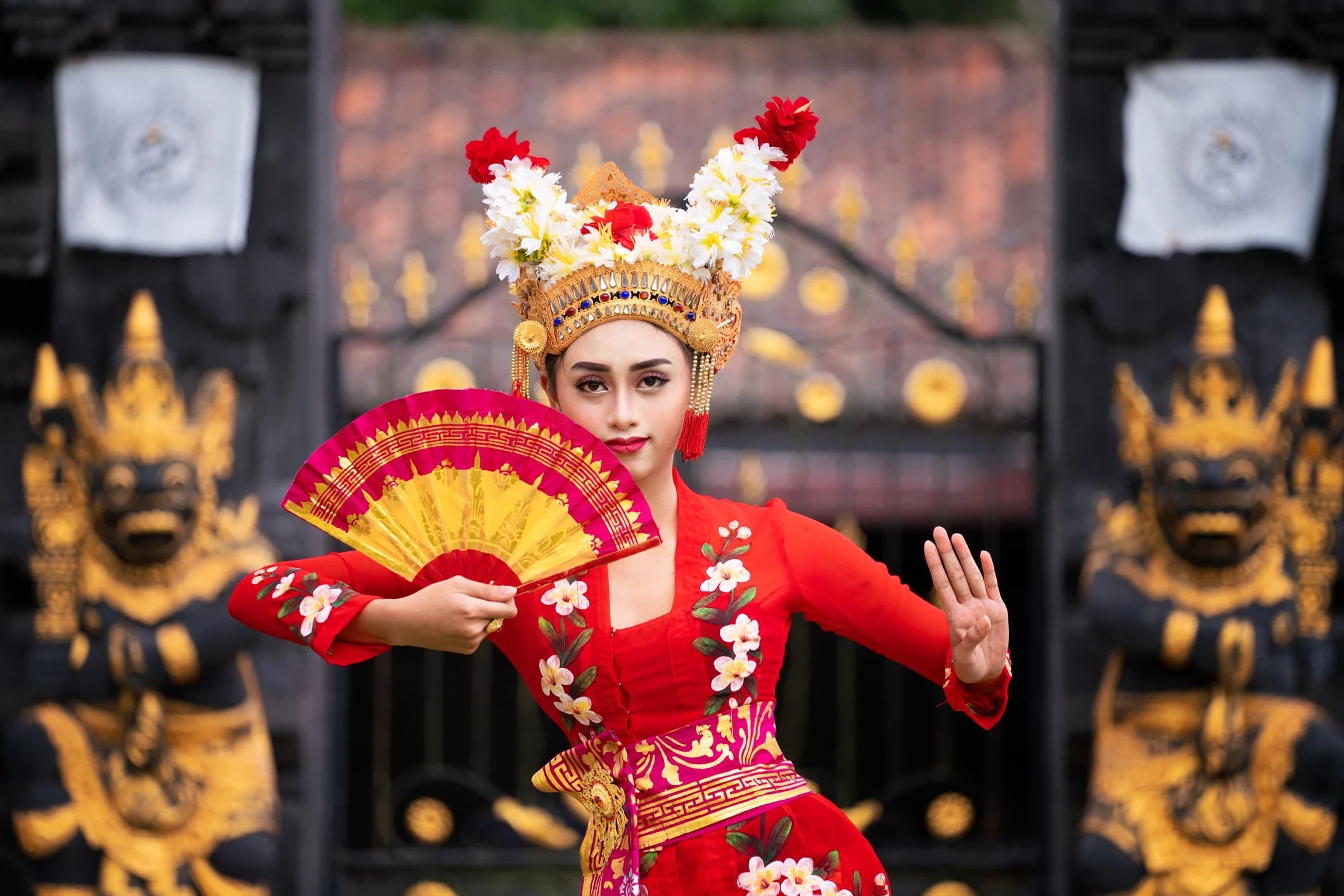 A woman in a vibrant red traditional dress holds a fan while posing in front of ornate temple gates.