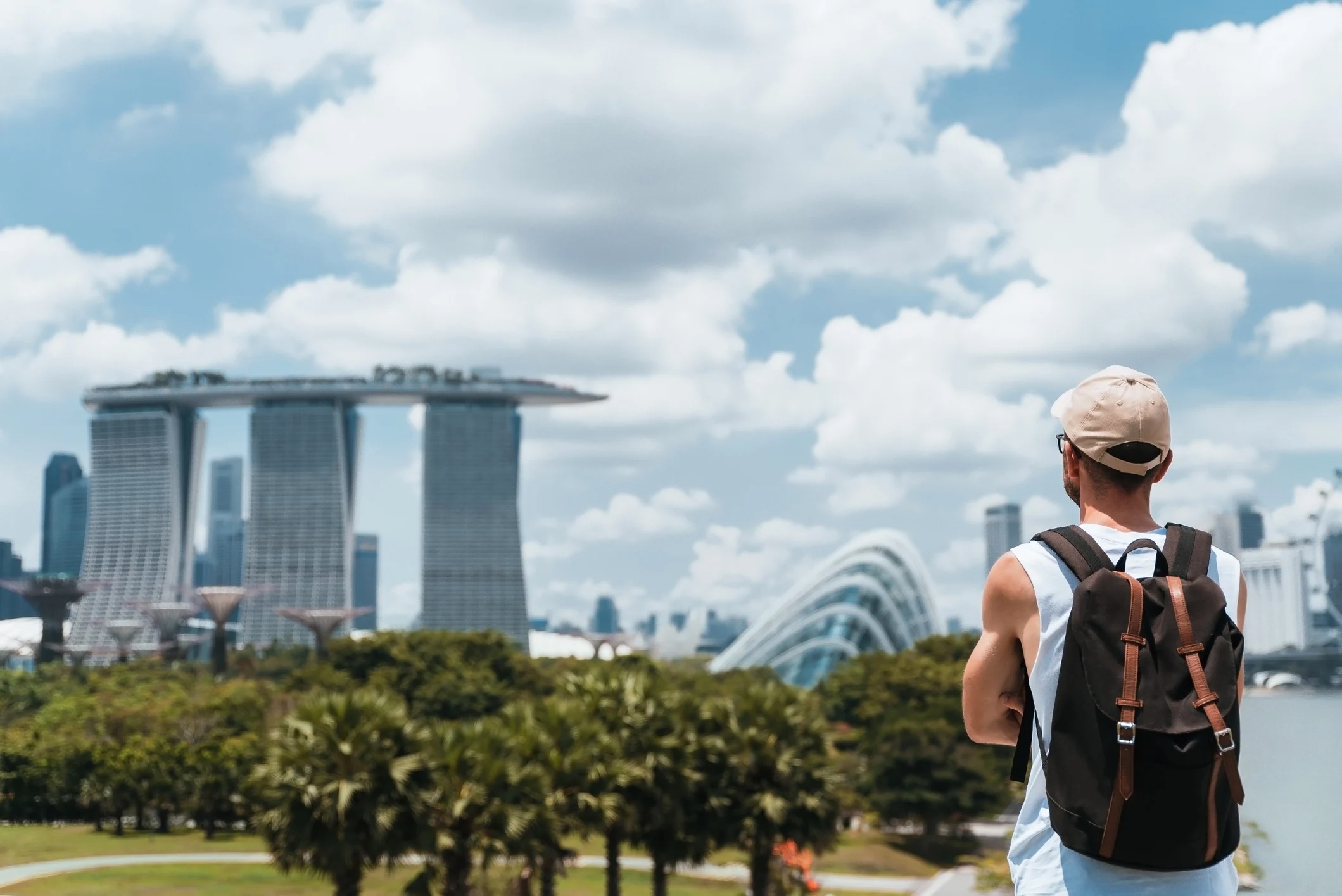 A person with a backpack gazes at futuristic buildings under a blue sky with clouds in a lush park setting.