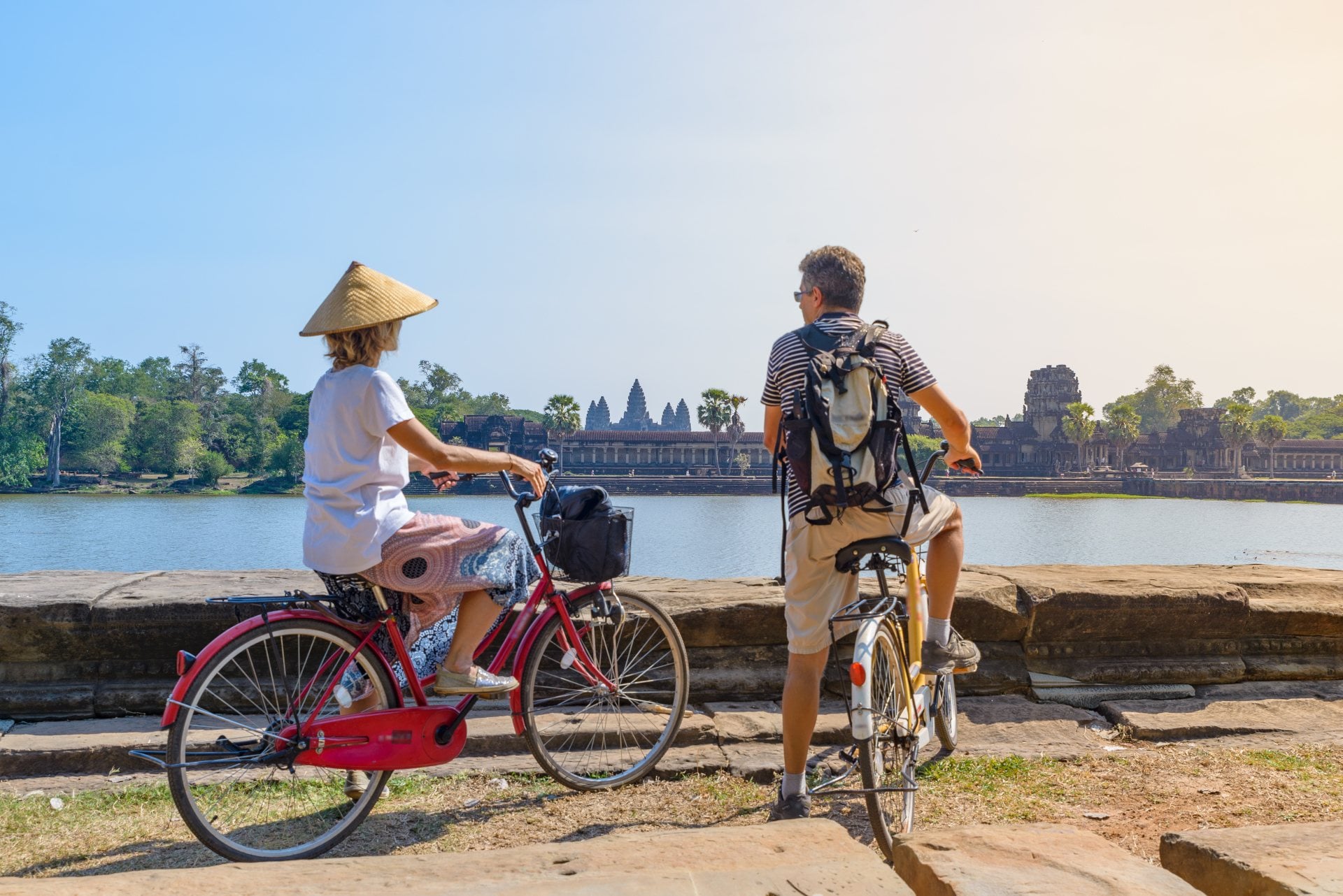 Two cyclists admire a historic temple by a lake in the background, enjoying a sunny day. One wears a traditional hat.