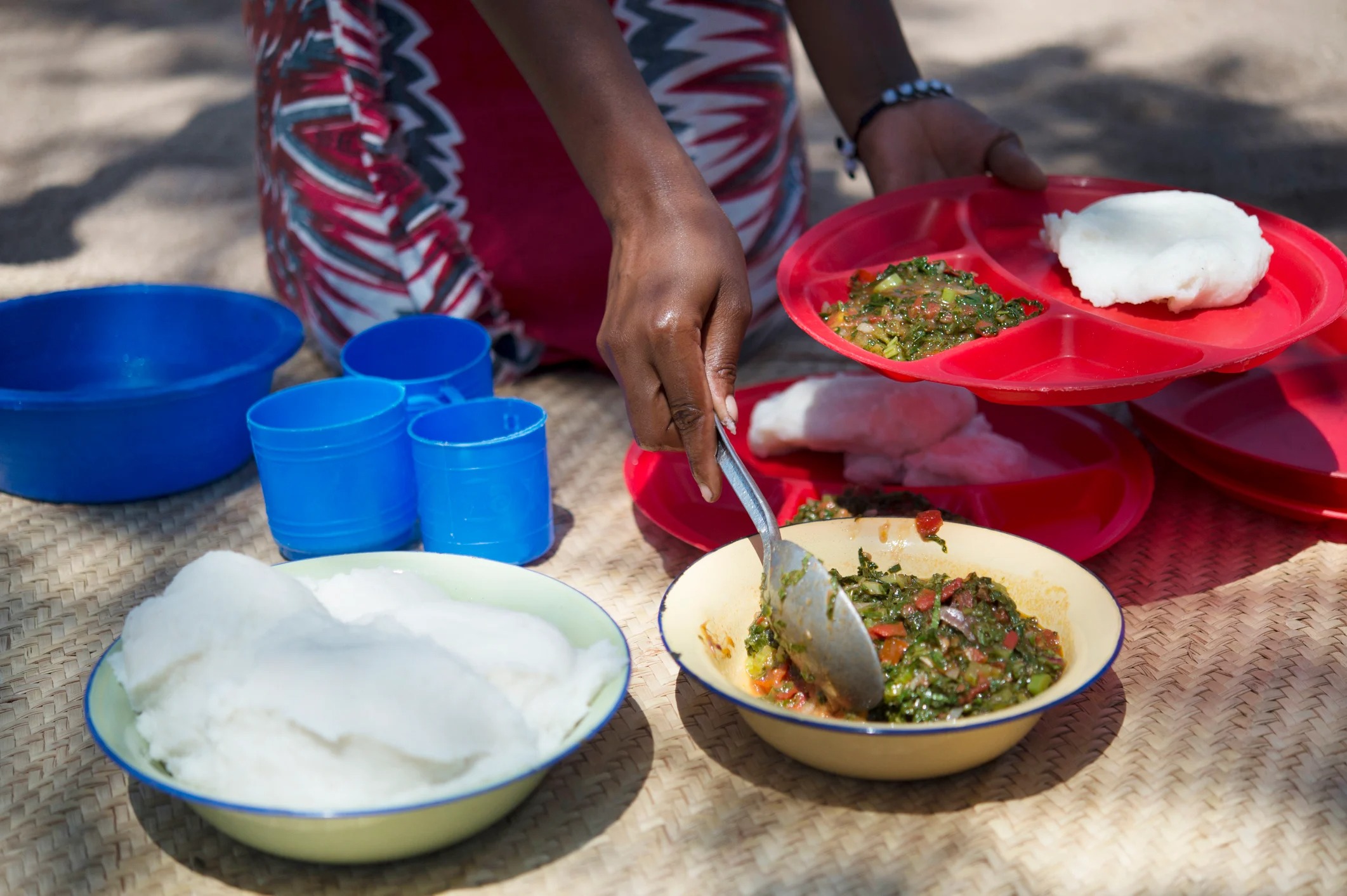 A person serves food from a bowl onto colorful plates, with cups nearby, on a textured surface outdoors.