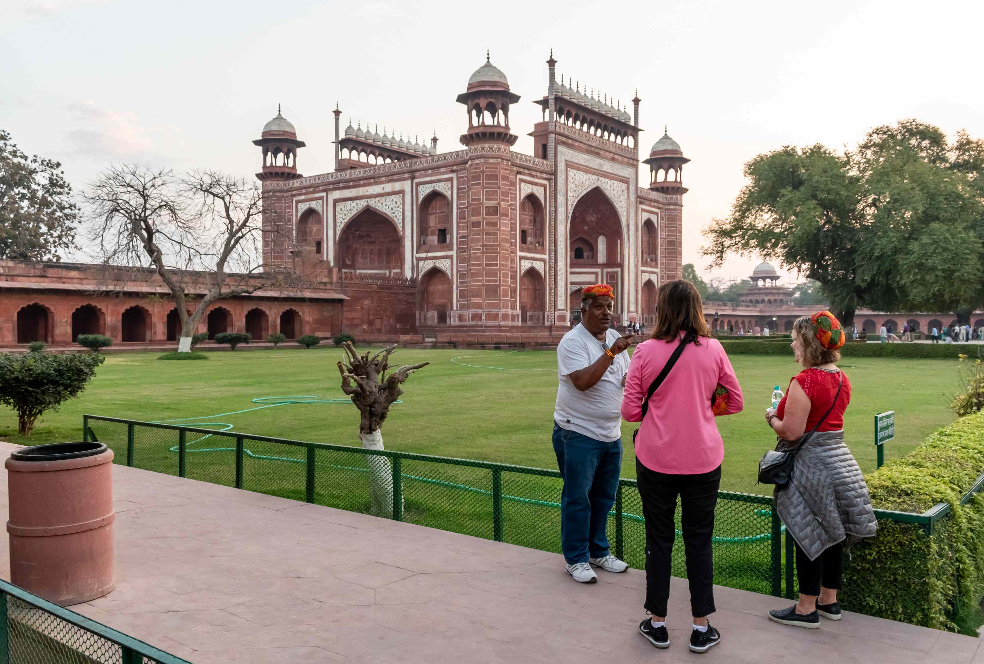 Visitors stand in a landscaped area, talking with an individual near a large red sandstone monument at sunset.