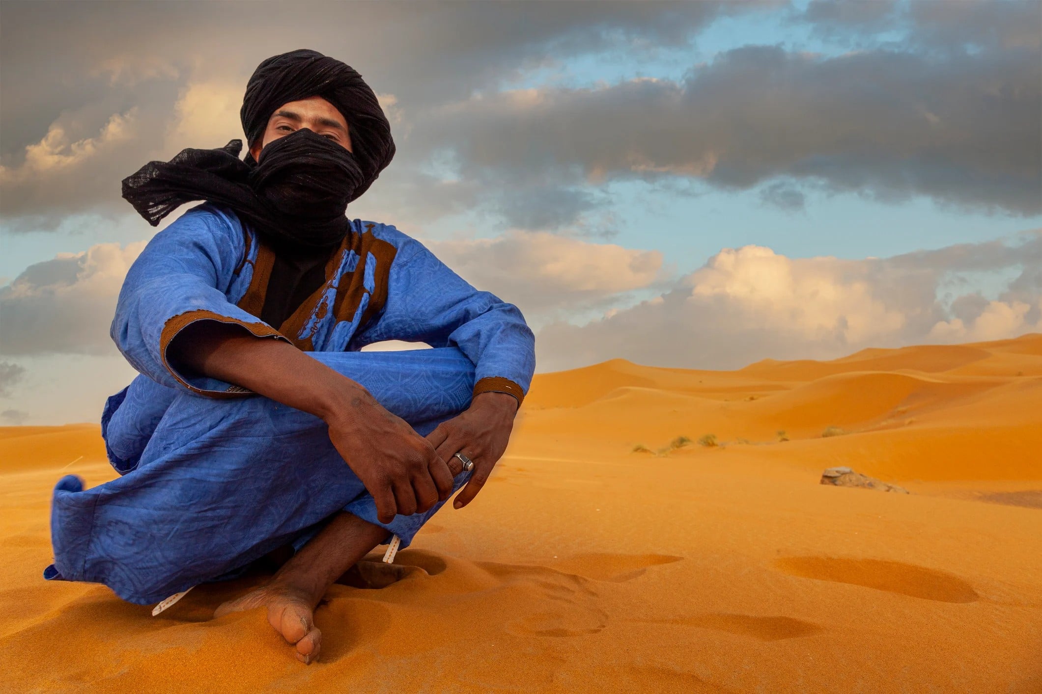 A person in a blue robe and black scarf sits on golden sand dunes under a cloudy sky.