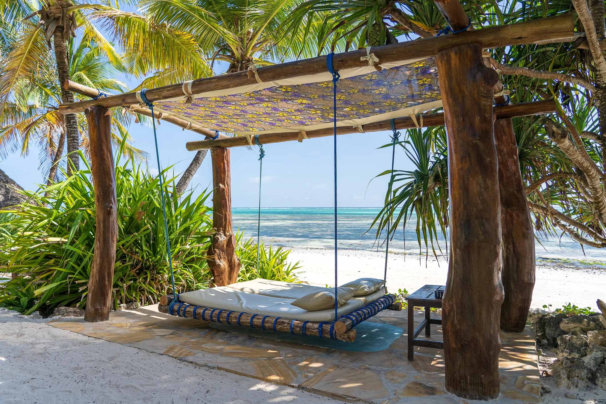 A shaded hammock swing under palm trees on a beach, with clear water and white sand in the background.