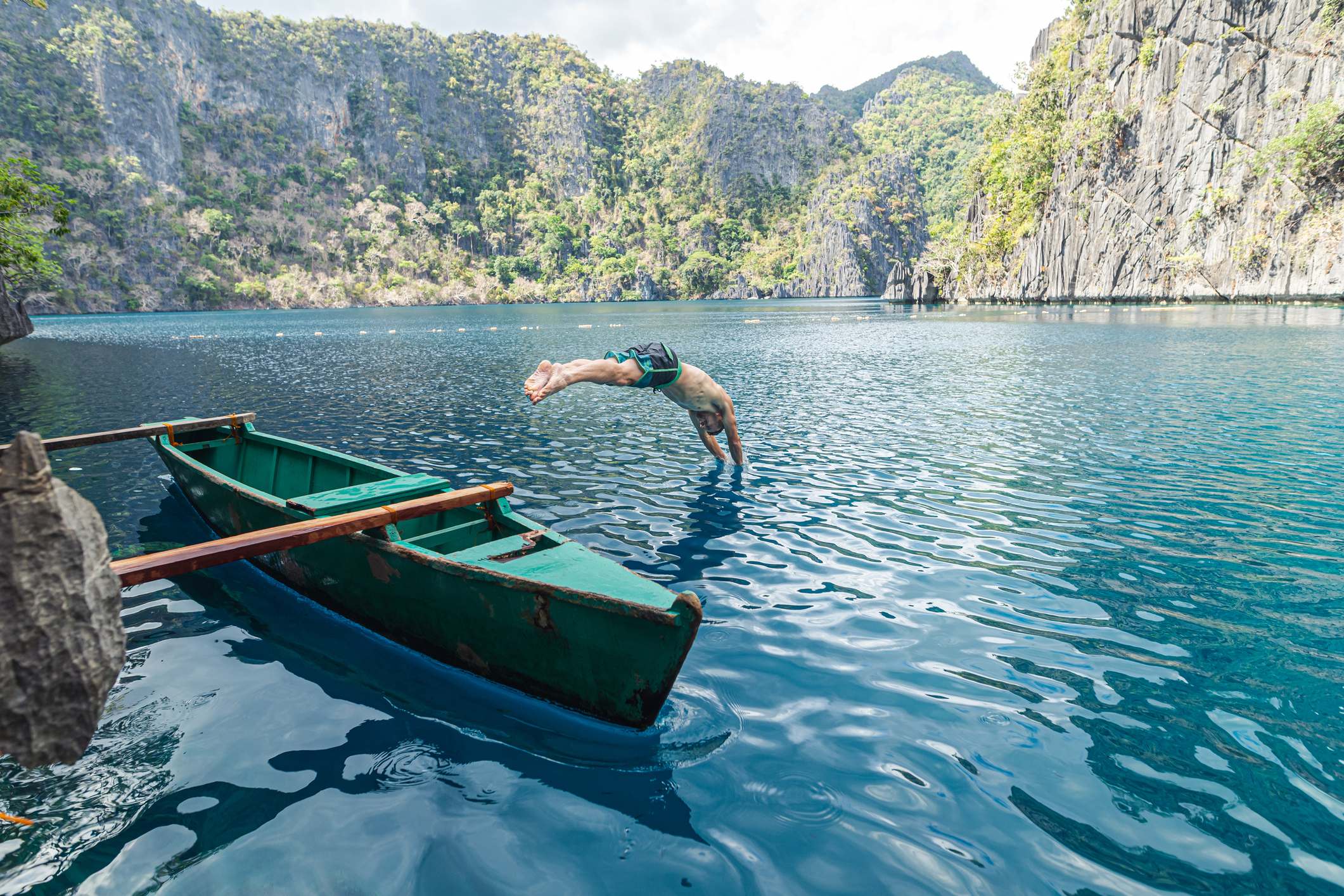 A person dives into clear blue water from the edge of a green boat surrounded by rocky cliffs and lush greenery.