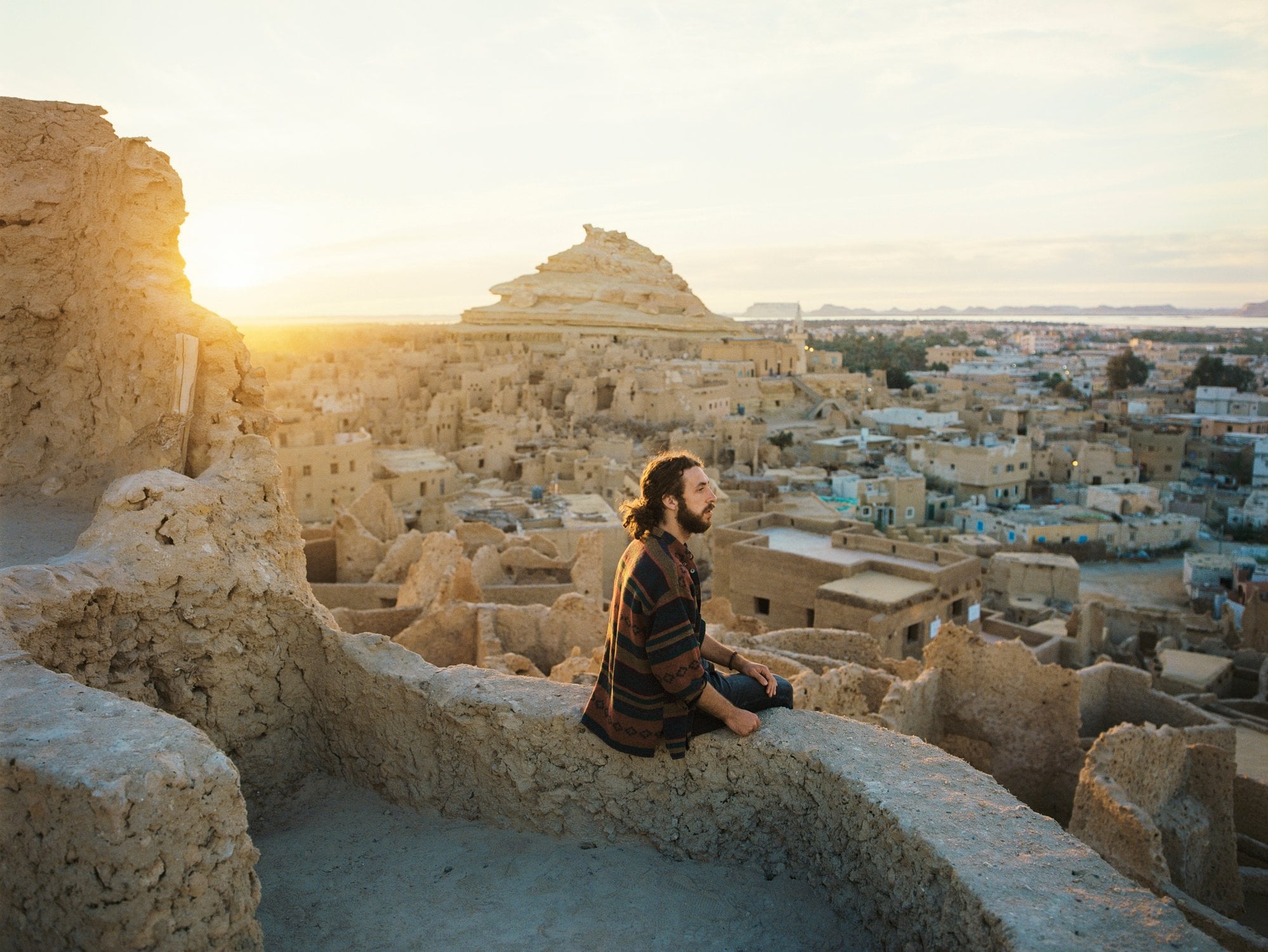A person sits on a wall overlooking a village at sunset, with ancient structures and a pyramid in the background.