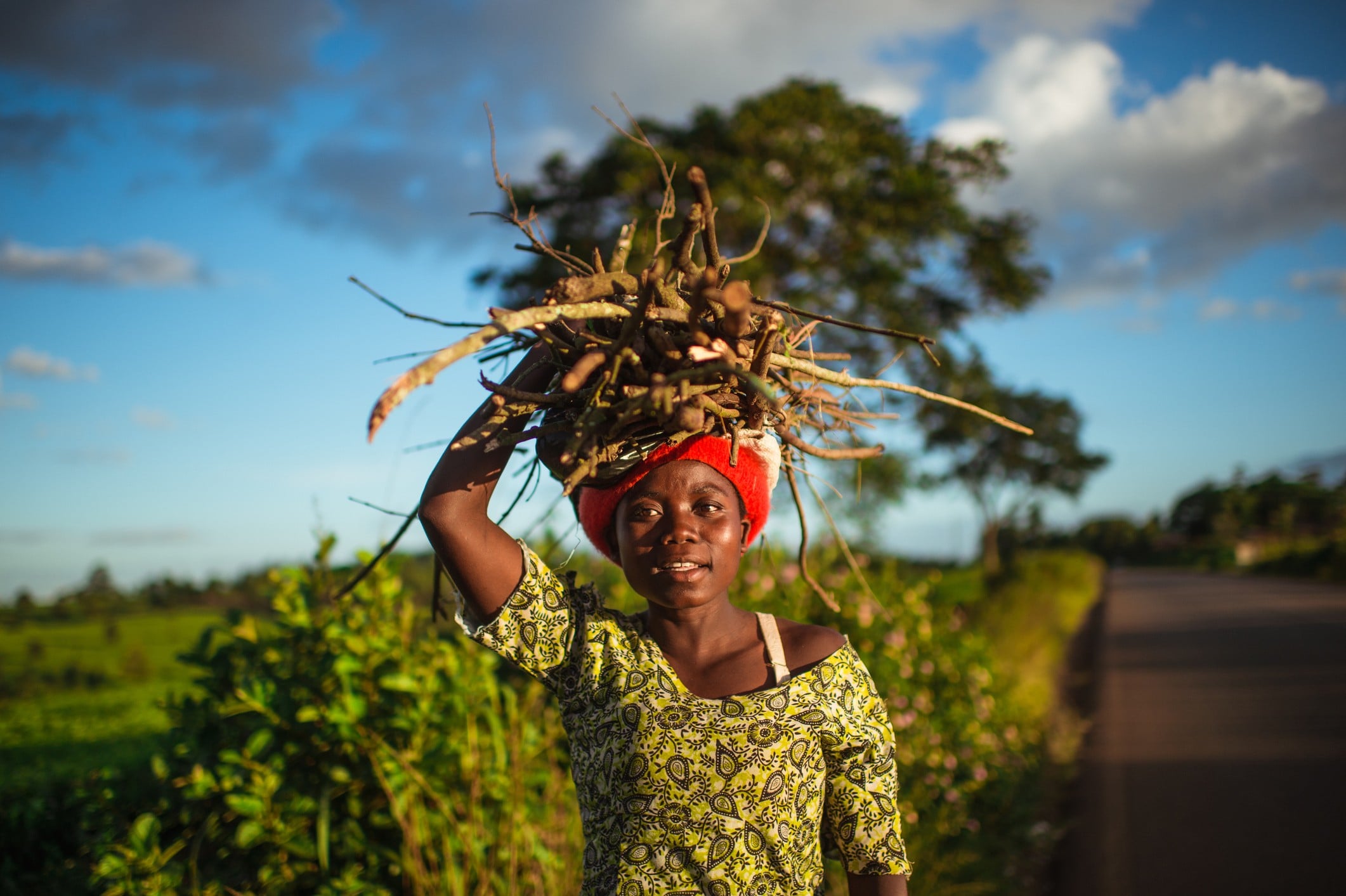 A girl with a red headwrap carries a large bundle of sticks on her head, standing on a rural road with greenery around.