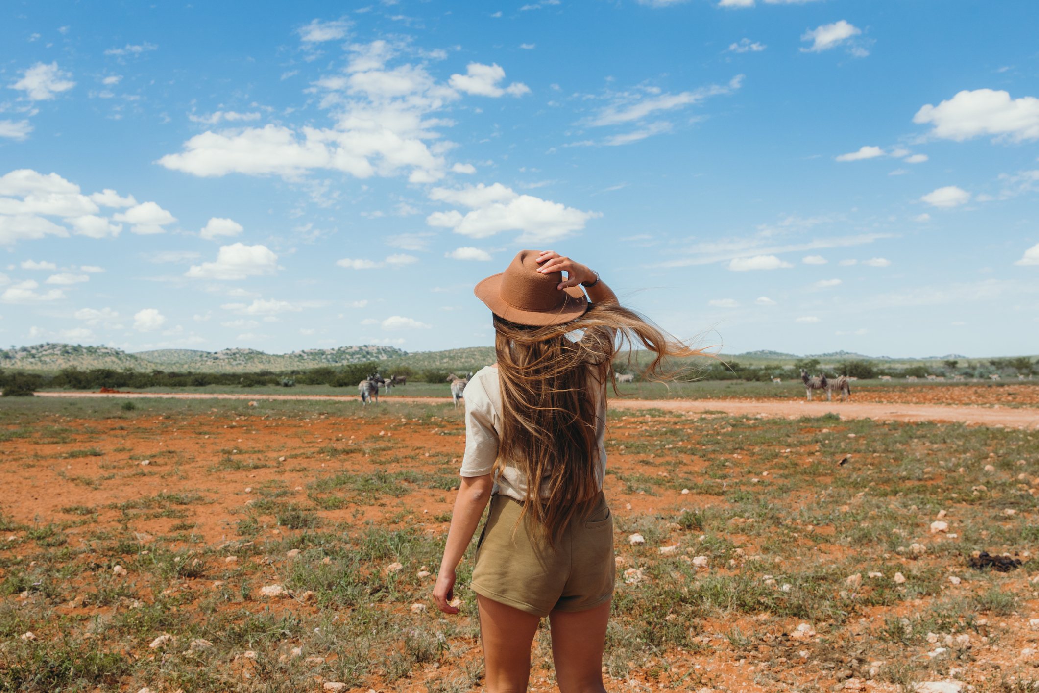 A person with long hair in a hat stands on a grassy plain, looking at a wide sky with scattered clouds.