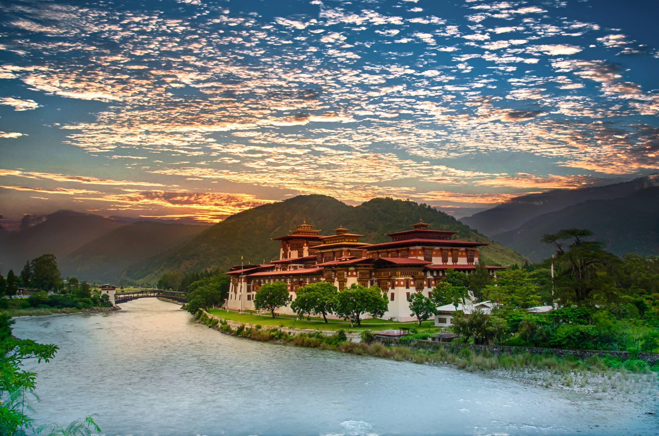 A scenic view of a traditional Bhutanese building by a river, surrounded by mountains and a vibrant sky at sunset.