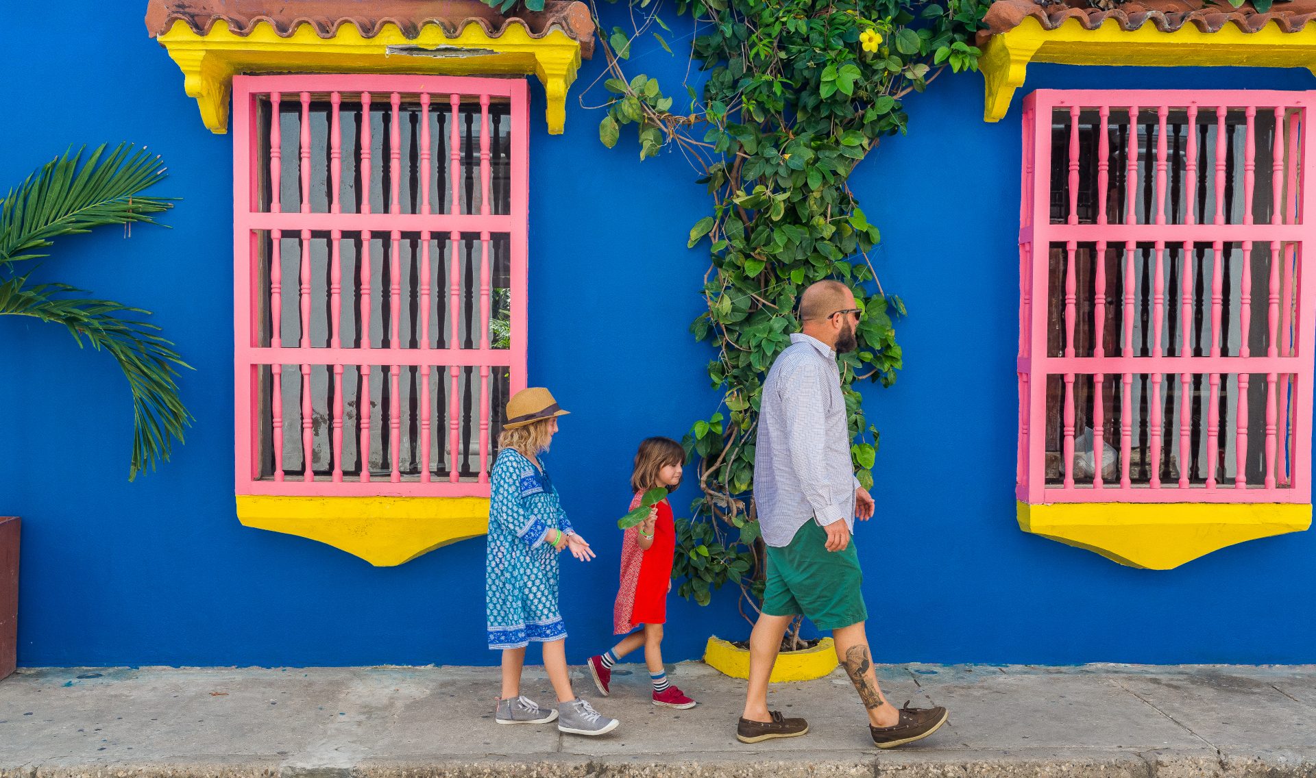 A family of four walks along a bright blue wall with pink windows and greenery, enjoying a sunny day.