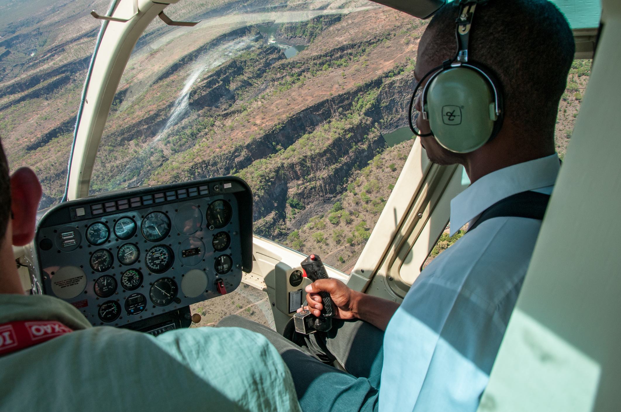 Pilot and passenger inside a helicopter, flying over a mountainous landscape, with cockpit instruments visible.