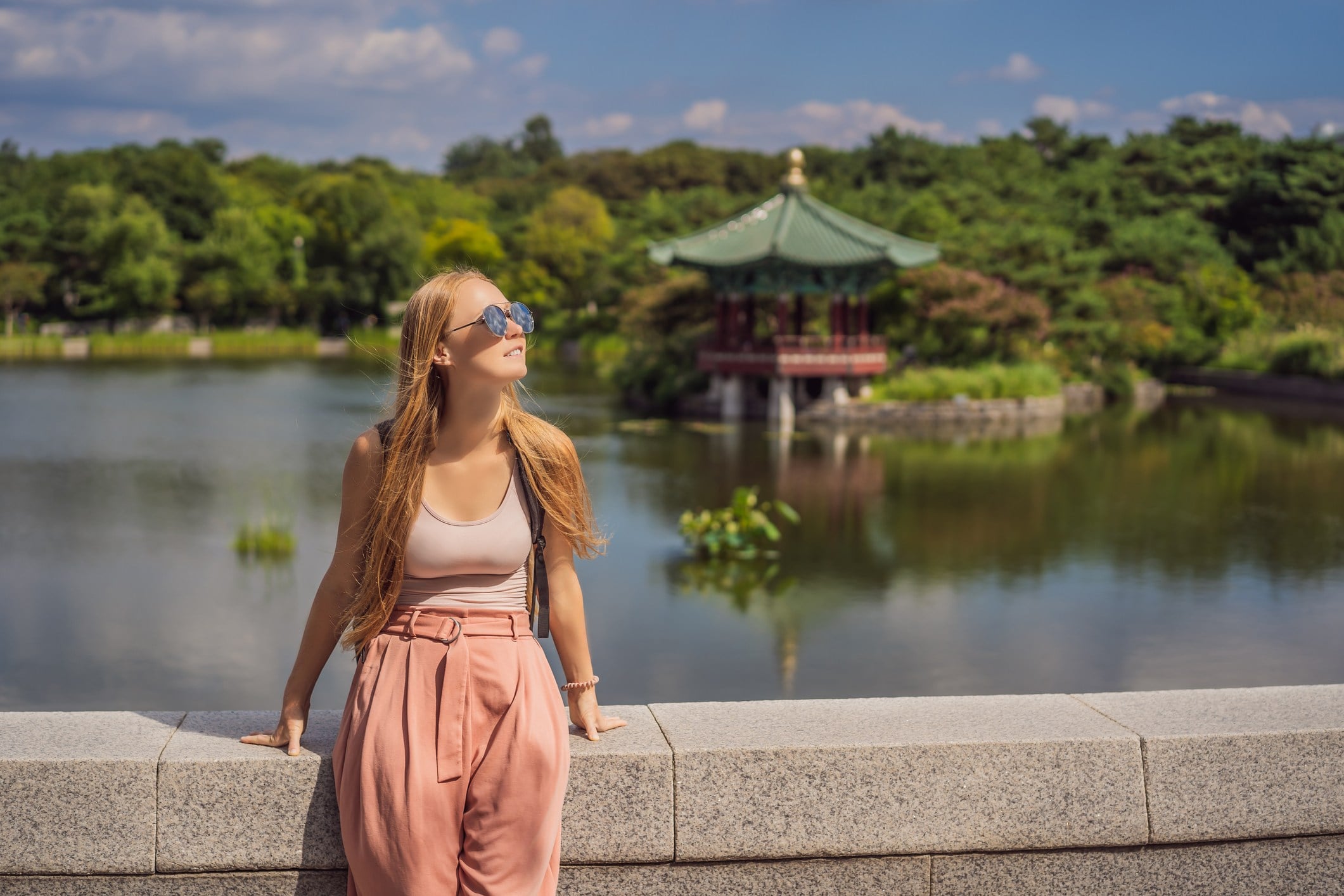 A young woman in sunglasses stands by a lake, smiling with a gazebo and lush greenery in the background.