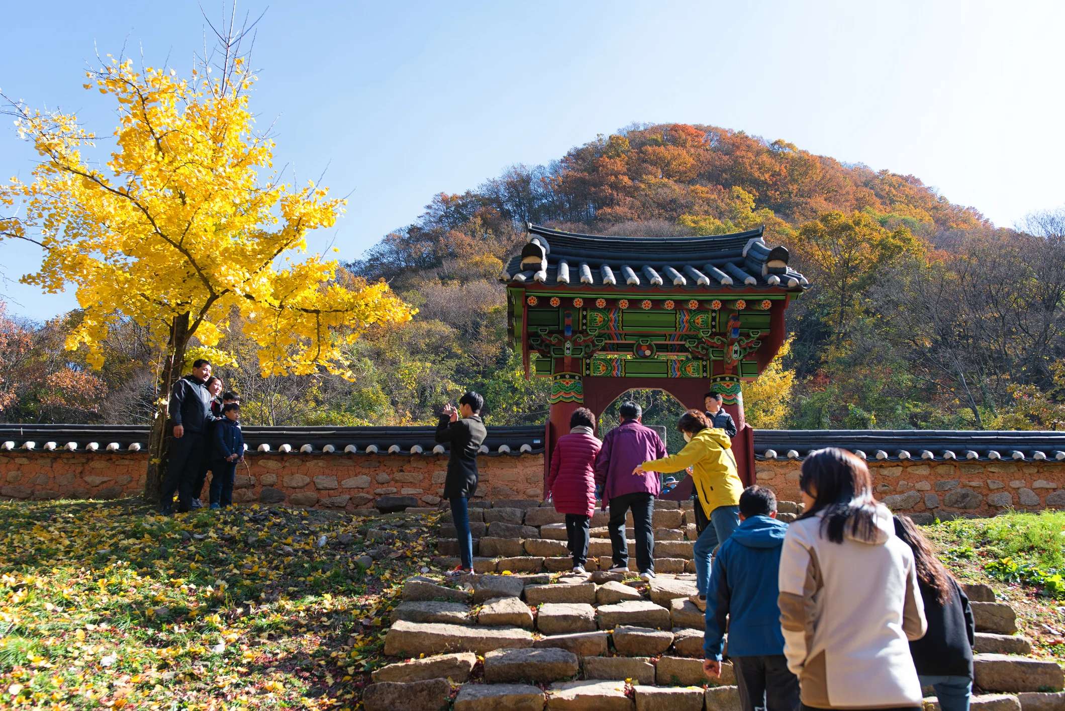 Visitors ascend stone steps toward a colorful traditional gate, surrounded by autumn trees and vibrant foliage.