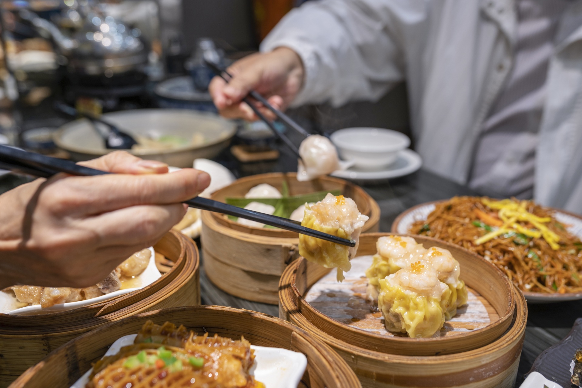 Hands using chopsticks to pick up dim sum from bamboo steamers on a table with noodles and other dishes in the background.