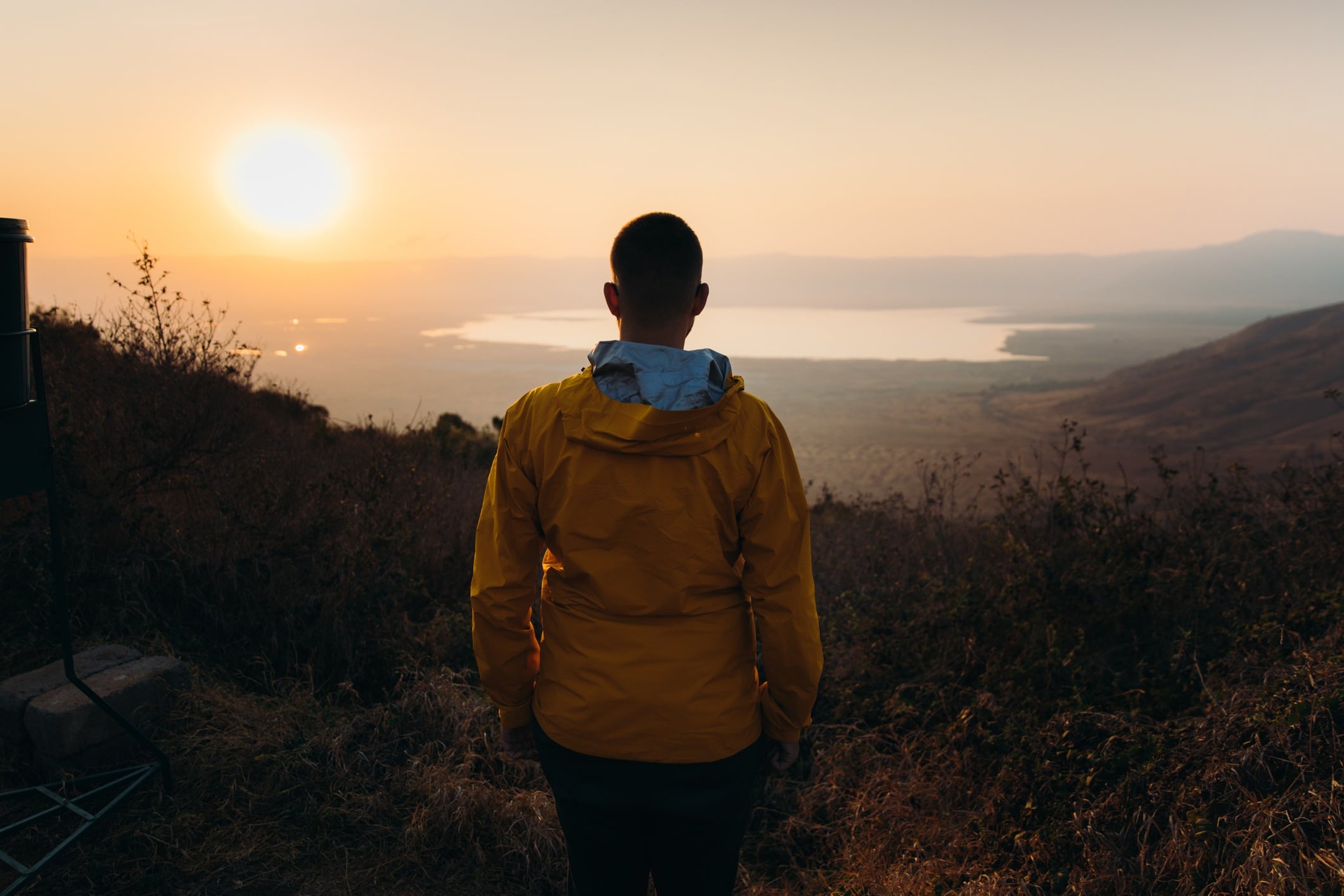A person in a yellow jacket stands facing a sunset over a lake, surrounded by hills and vegetation.