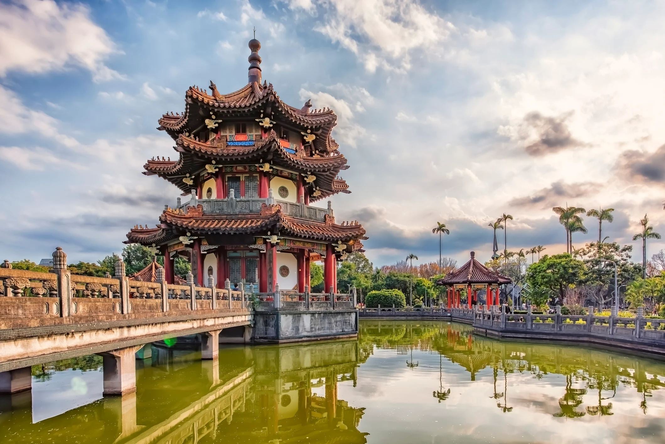 A traditional pagoda beside a tranquil pond, surrounded by lush greenery and a cloudy sky.