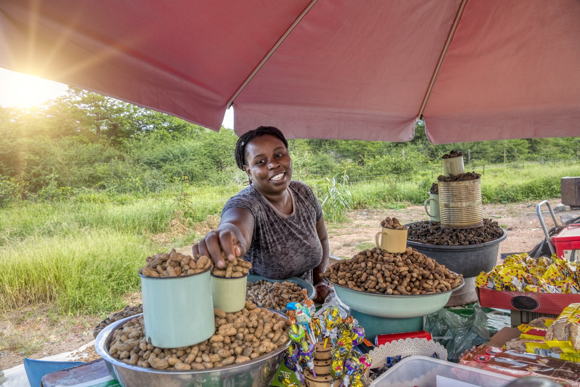 A smiling vendor at a market offers a cup of peanuts, surrounded by various snacks under a shaded tent.