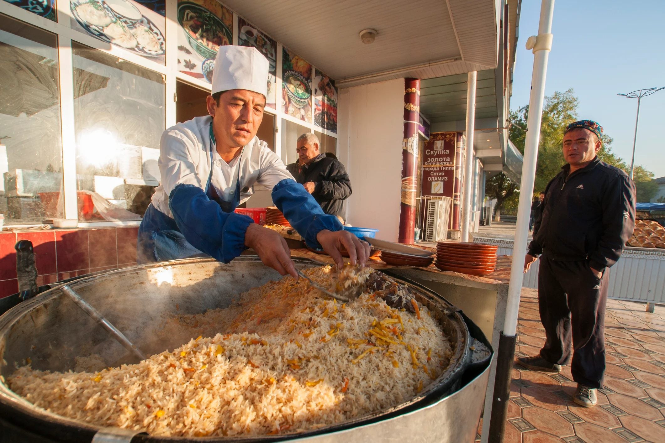 A chef in a white hat prepares a large pot of rice dish outdoors, while a man waits nearby.