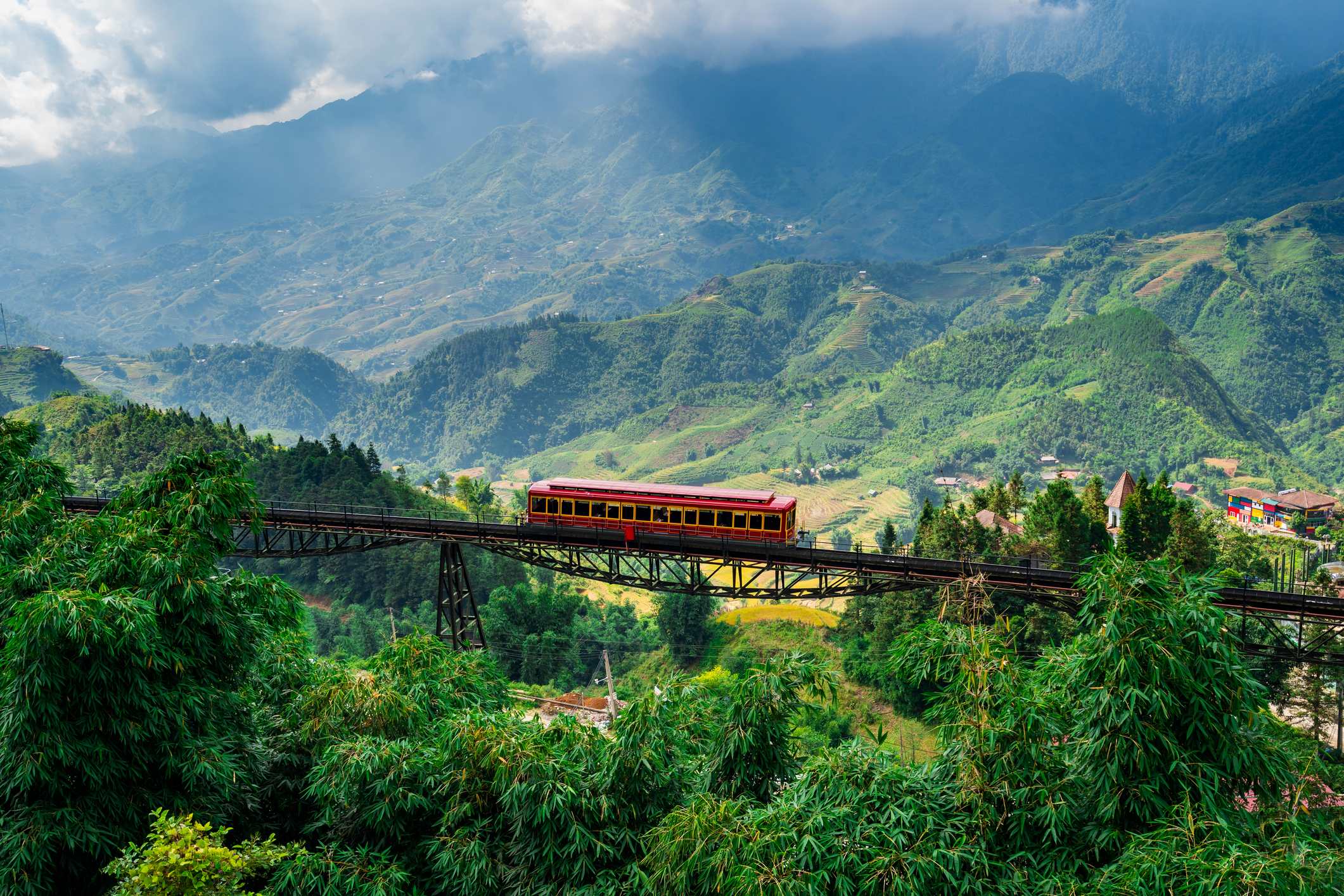 A red train crosses a bridge over lush green mountains under a cloudy sky.