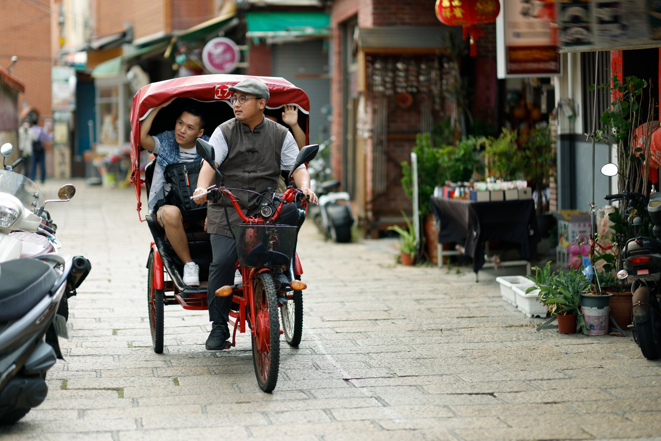 A man rides a pedicab with a child along a narrow street lined with shops and greenery.