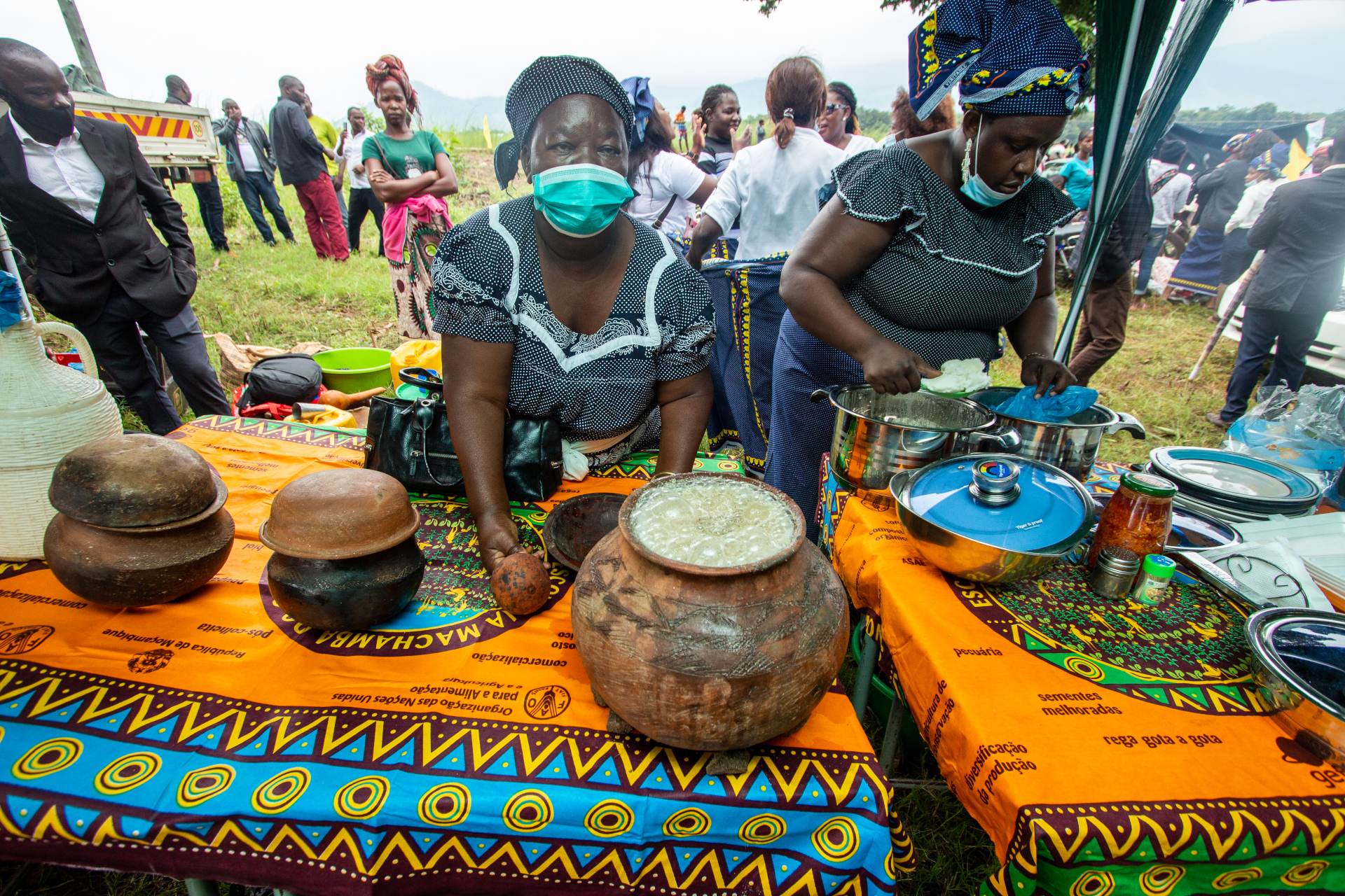Two women prepare food outdoors at a vibrant market, with traditional dishes displayed on colorful patterned tablecloths.