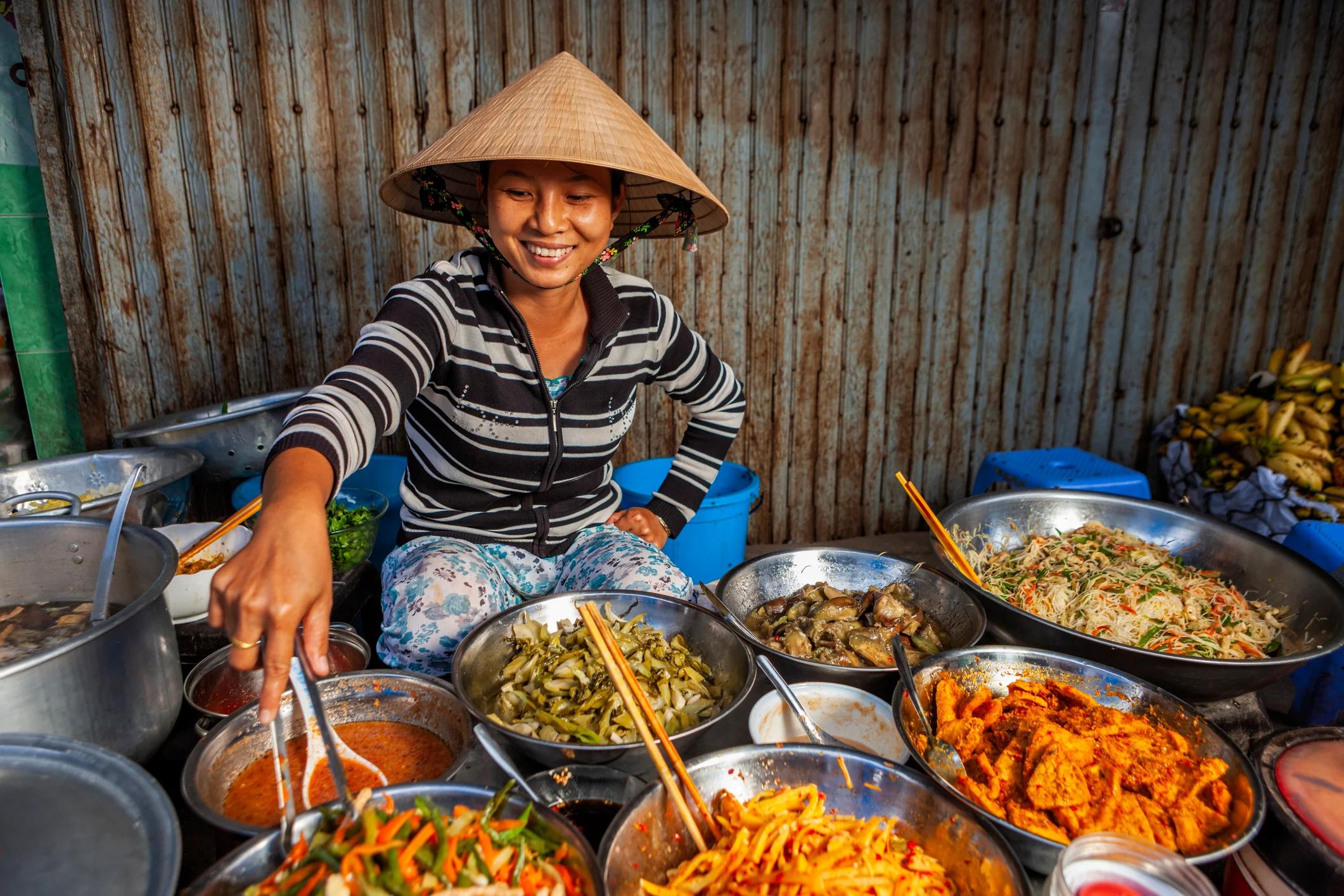 A smiling woman in a conical hat serves dishes from a variety of bowls at a vibrant market stall.